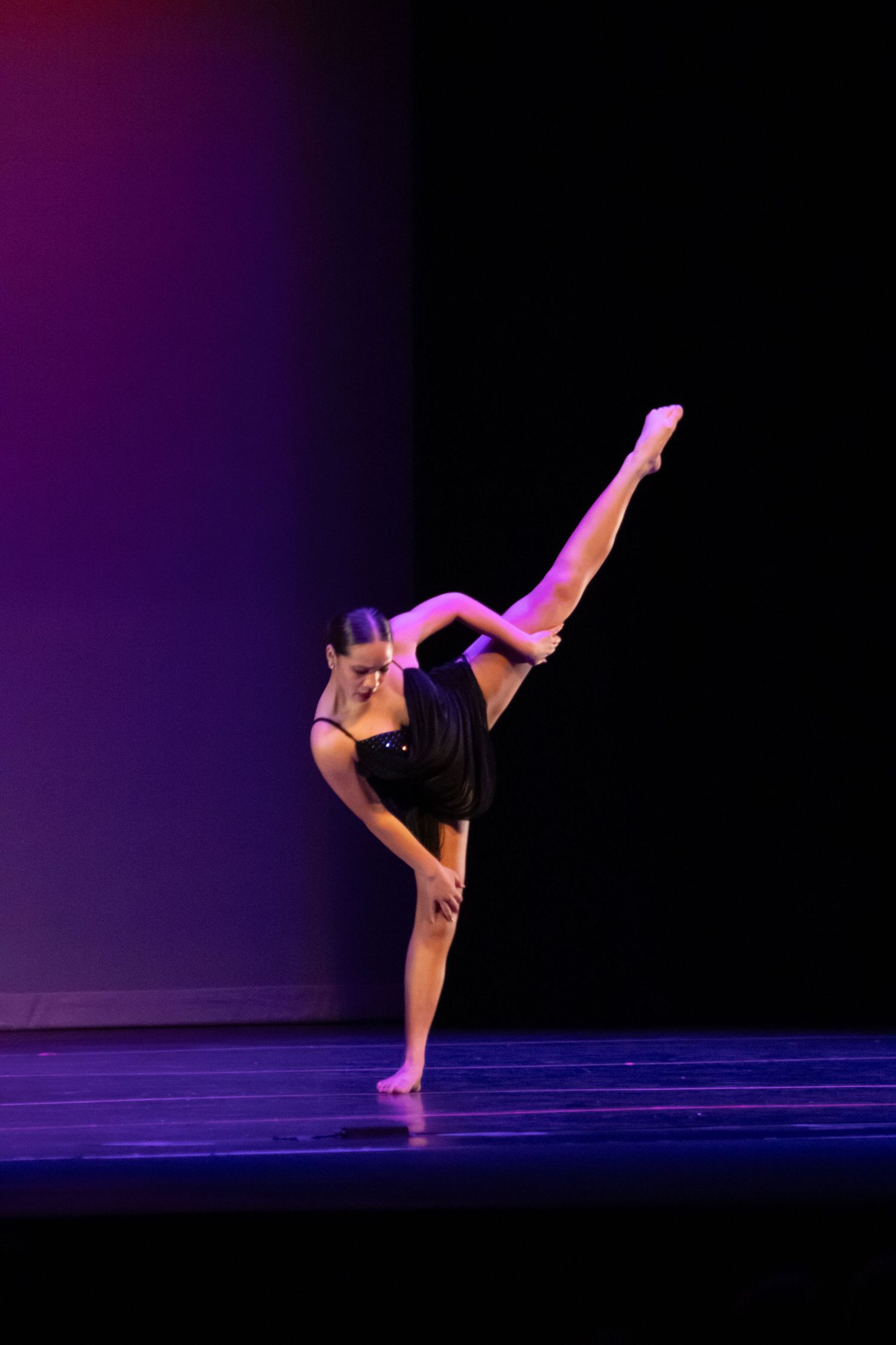 A dancer in black performs a high leg extension on a purple and blue lit stage.