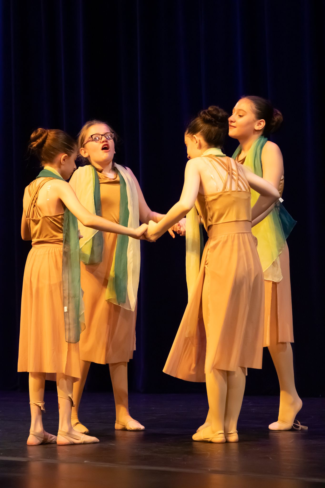 Four young girls in peach dresses and green scarves hold hands in a circle on stage.