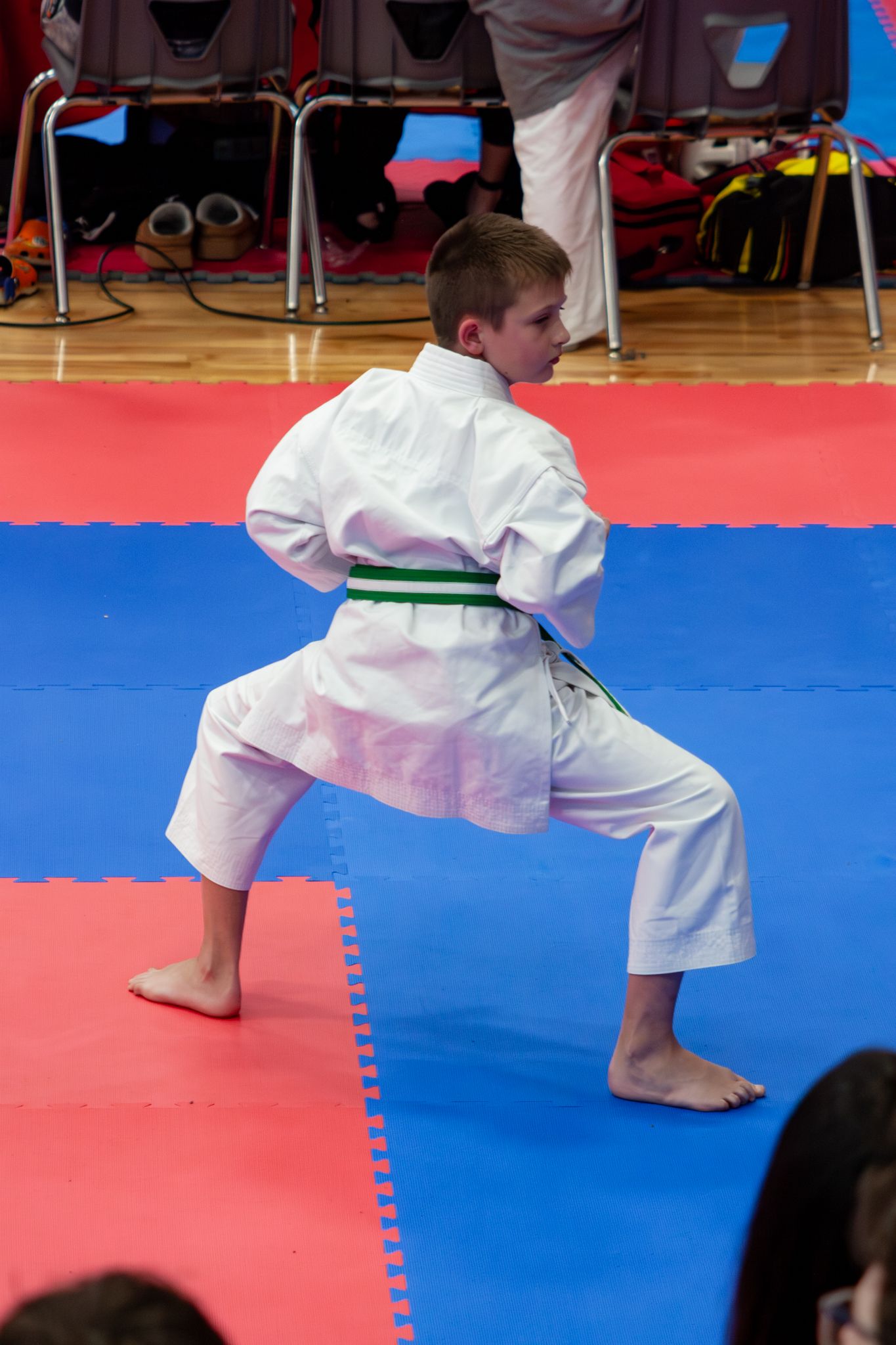 A barefoot child in a white gi and green belt practices a karate stance on colorful competition mats.