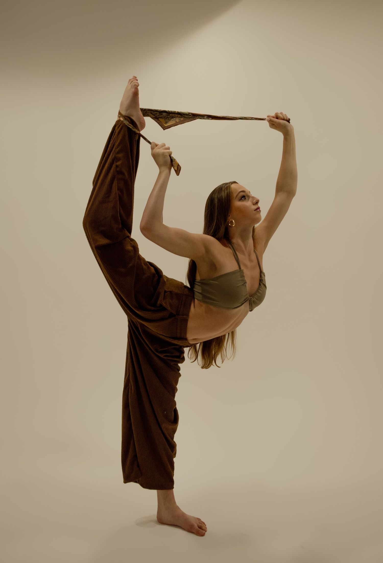 Flexible dancer balances on one leg in brown wide-leg pants and olive crop top against neutral background.