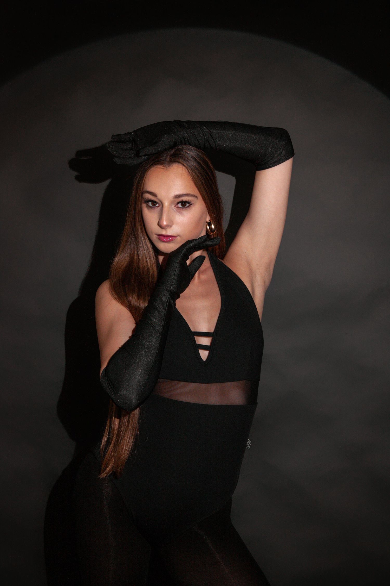 Young woman in black bodysuit and long black gloves poses elegantly against dark studio background.
