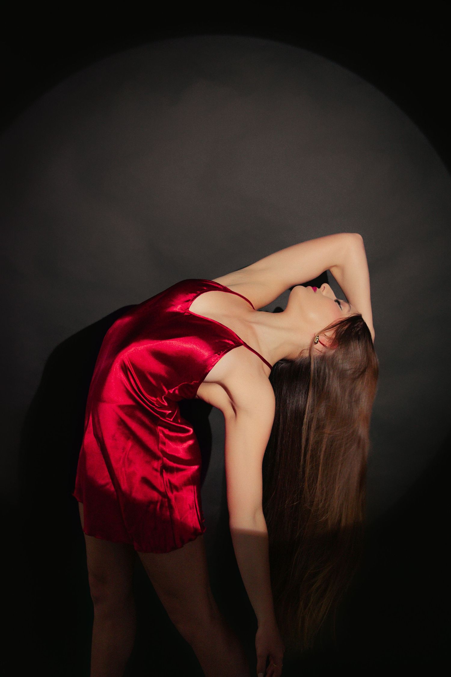 Woman in red satin mini dress arches back gracefully against dark studio spotlight background.