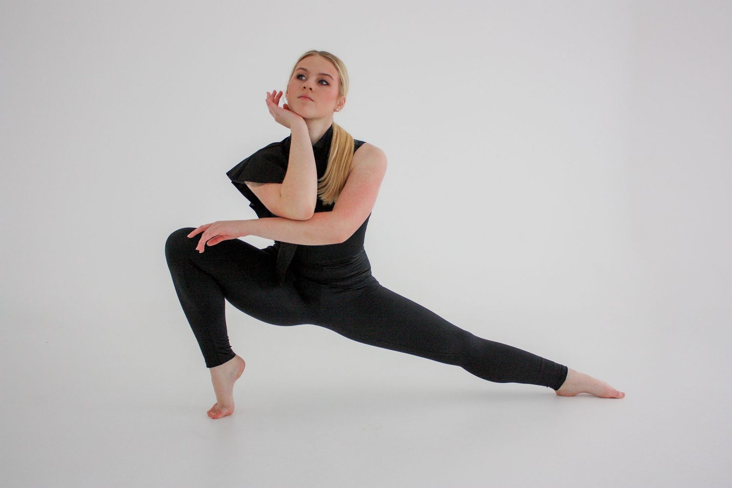 Blonde dancer in black attire holds deep lunge pose on white studio background.