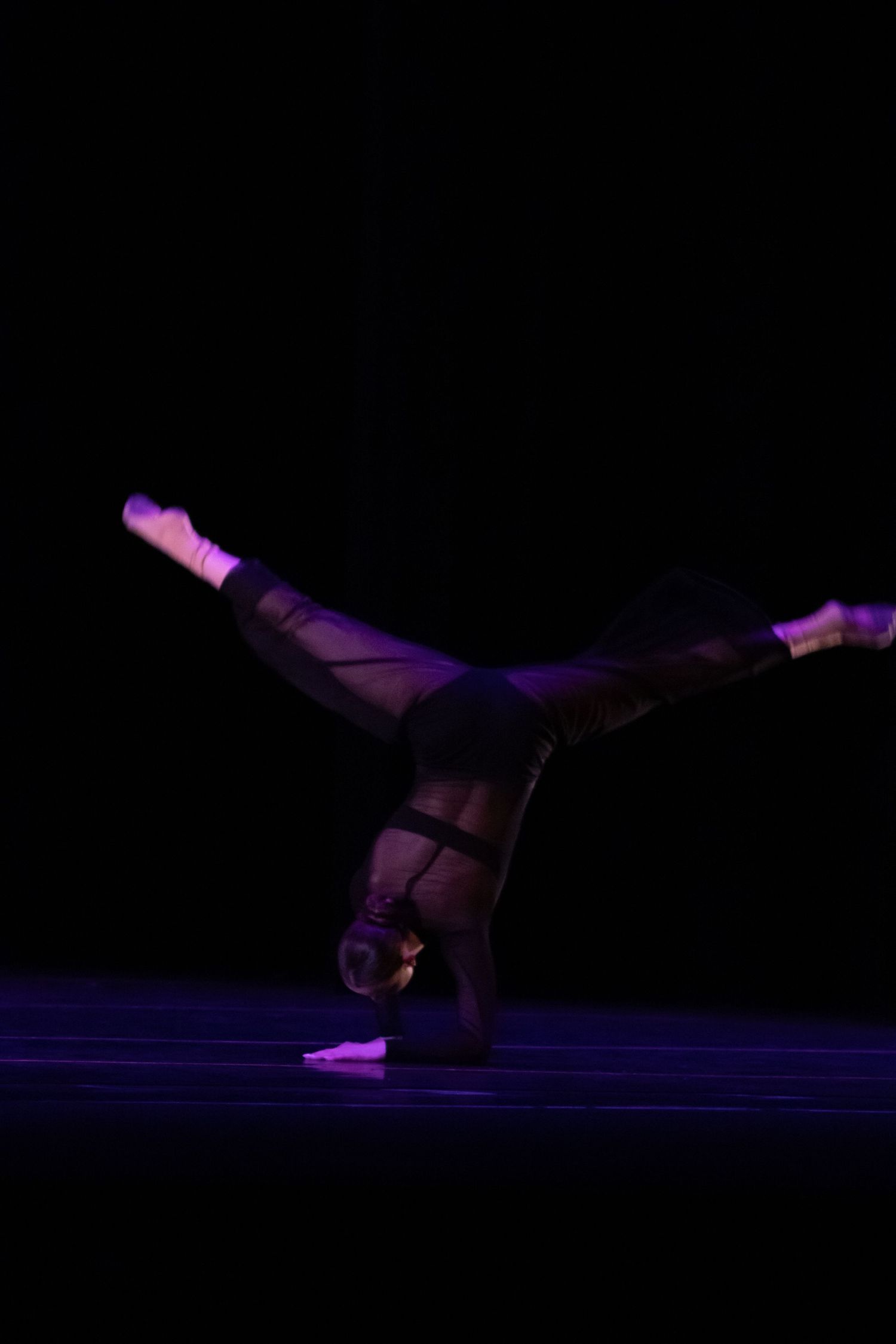 A dancer balances in a dramatic one-armed handstand with legs split on a dark stage.
