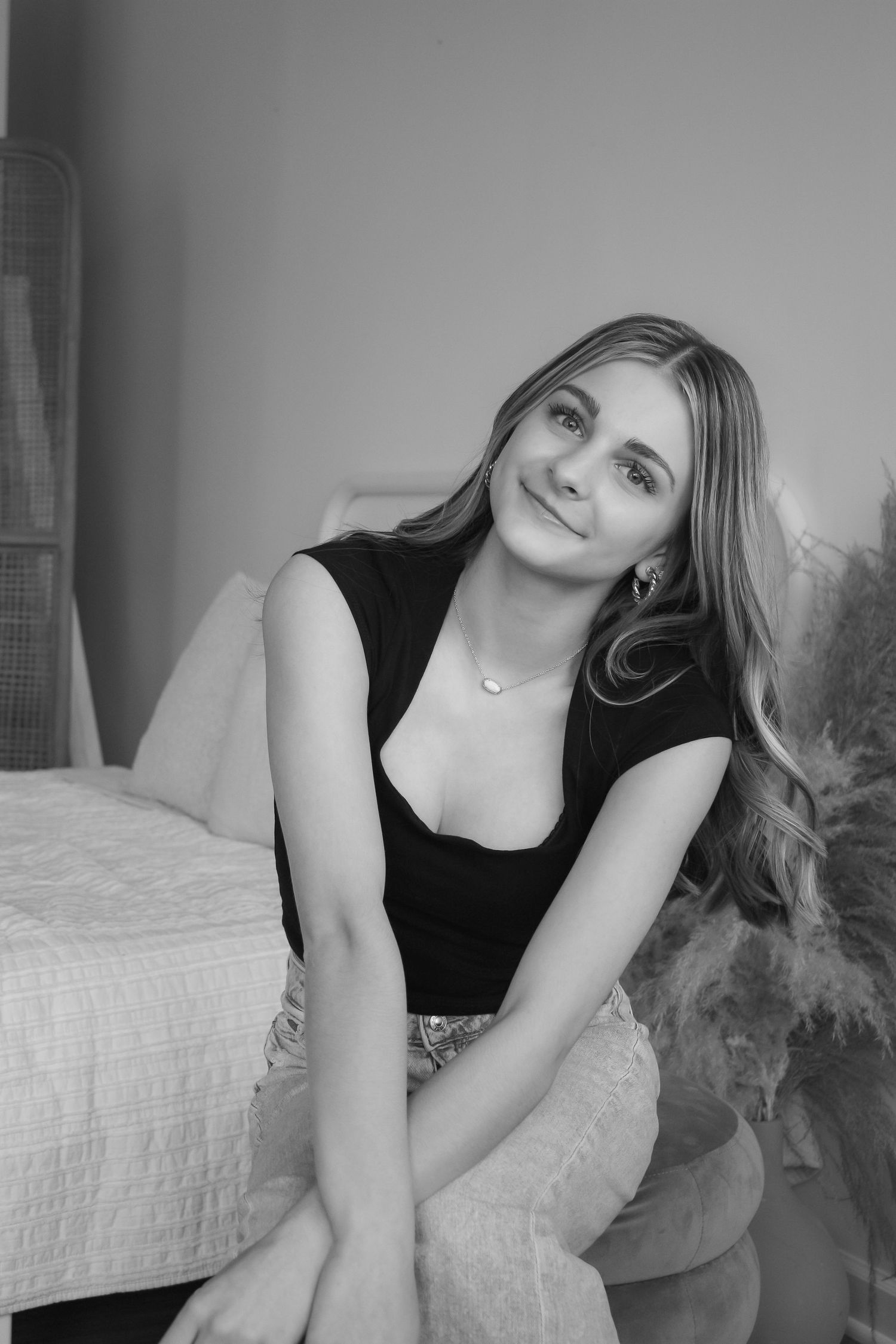 Black and white portrait of a young woman with long wavy hair sitting relaxed, wearing a black top and necklace.