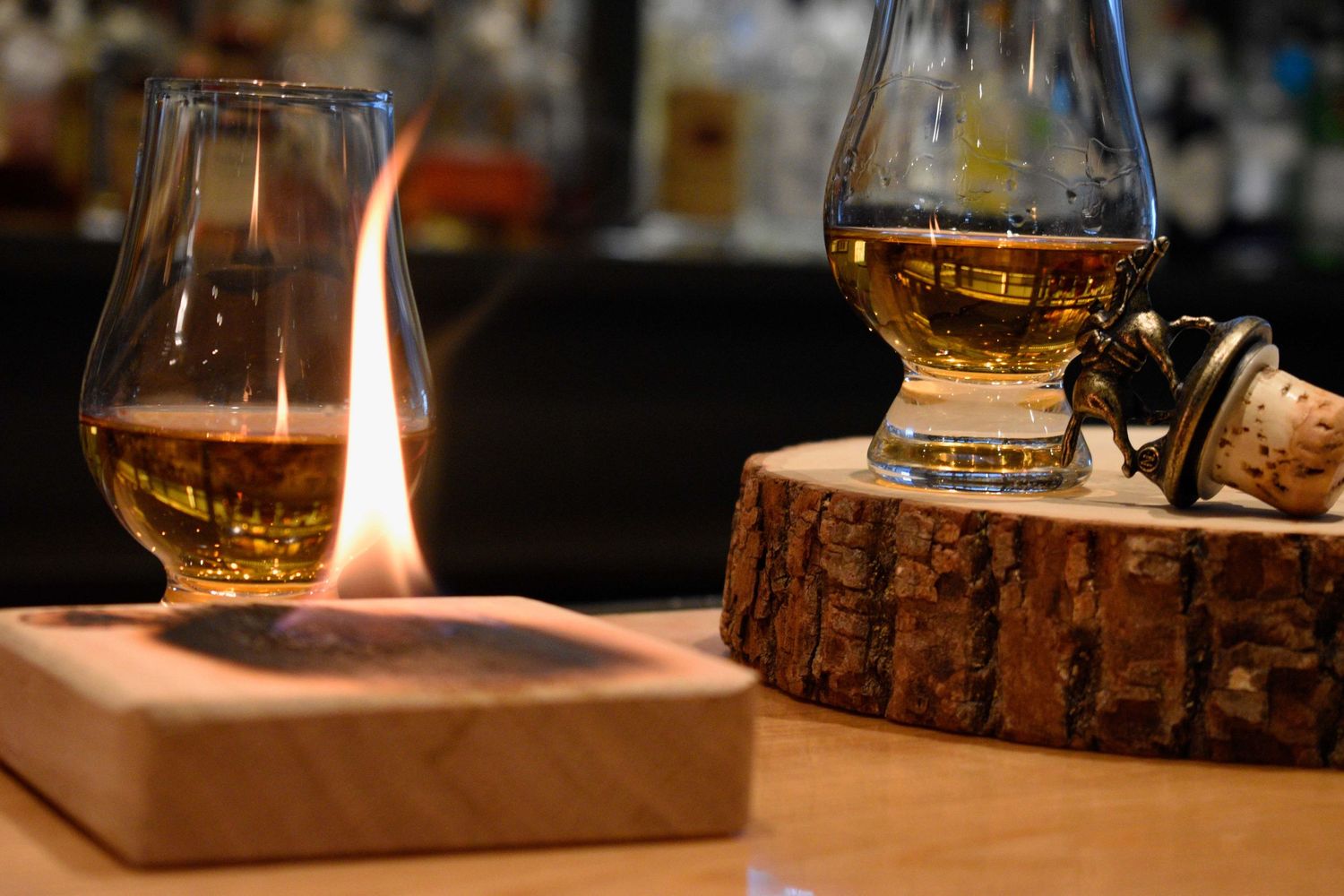 Whisky glass with a flame on a wooden coaster beside a Glencairn glass at a bar.