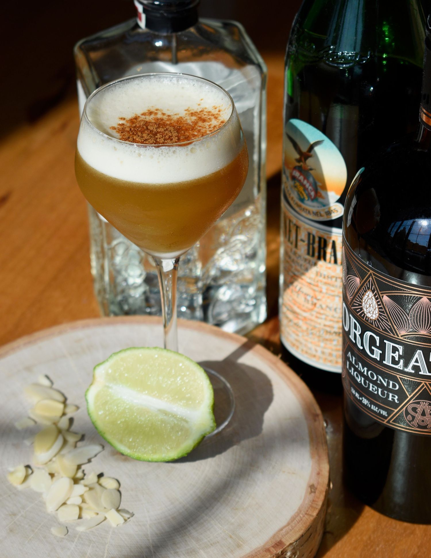 Frothy pisco sour cocktail with cinnamon on a wooden board beside Orgeat almond liqueur and tequila bottles.