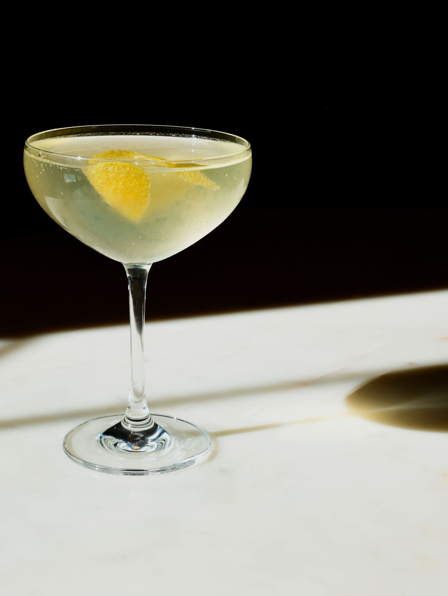 A classic coupe glass with a clear pale cocktail and lemon peel garnish, set against a dramatic dark background.