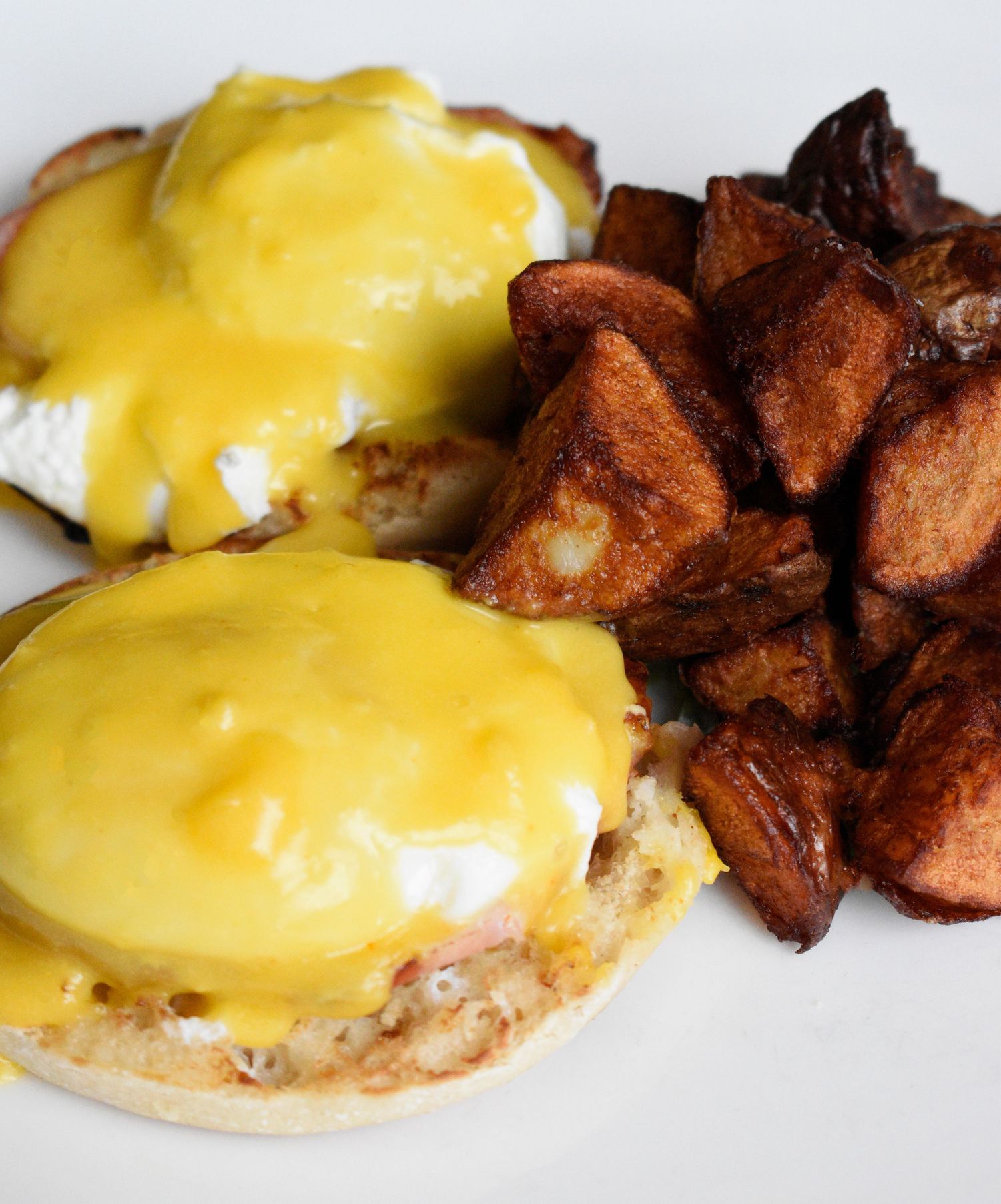 Classic eggs Benedict with hollandaise sauce served alongside crispy roasted potato cubes on a white plate.