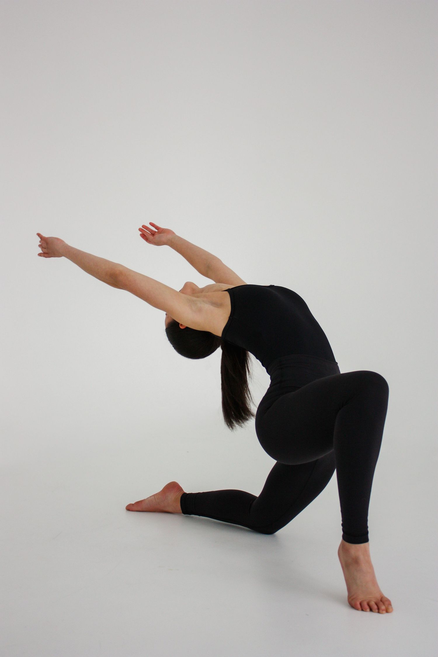 A female dancer in a black outfit performs a deep lunge with arms stretched forward against a white background.