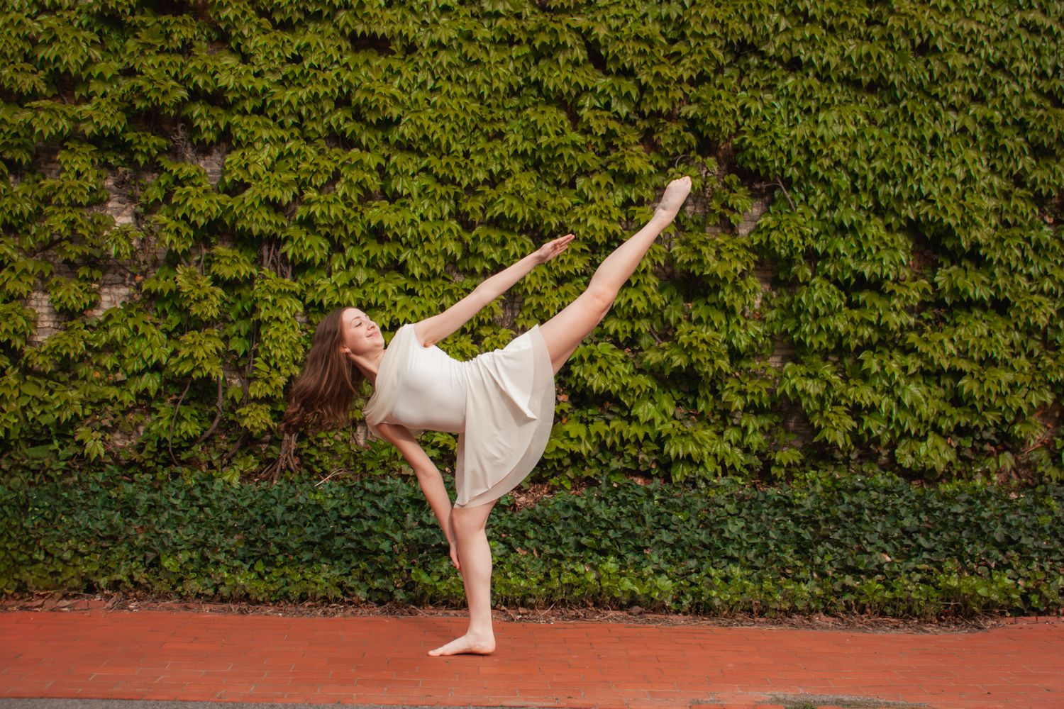 A barefoot dancer in a white dress executes a high kick balance pose in front of a green ivy-covered wall.