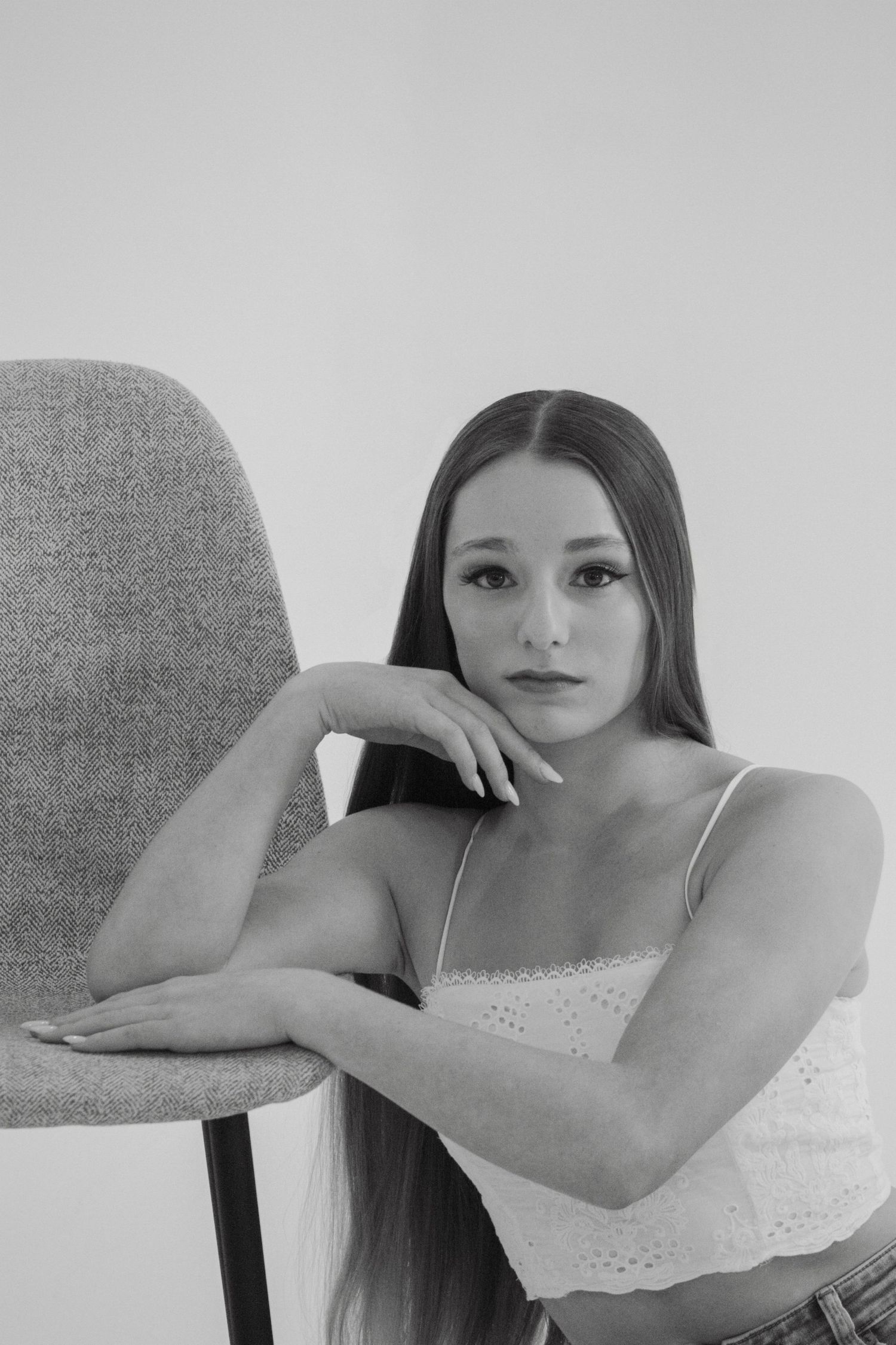 Young woman in white camisole leans on chair in soft black and white studio portrait.