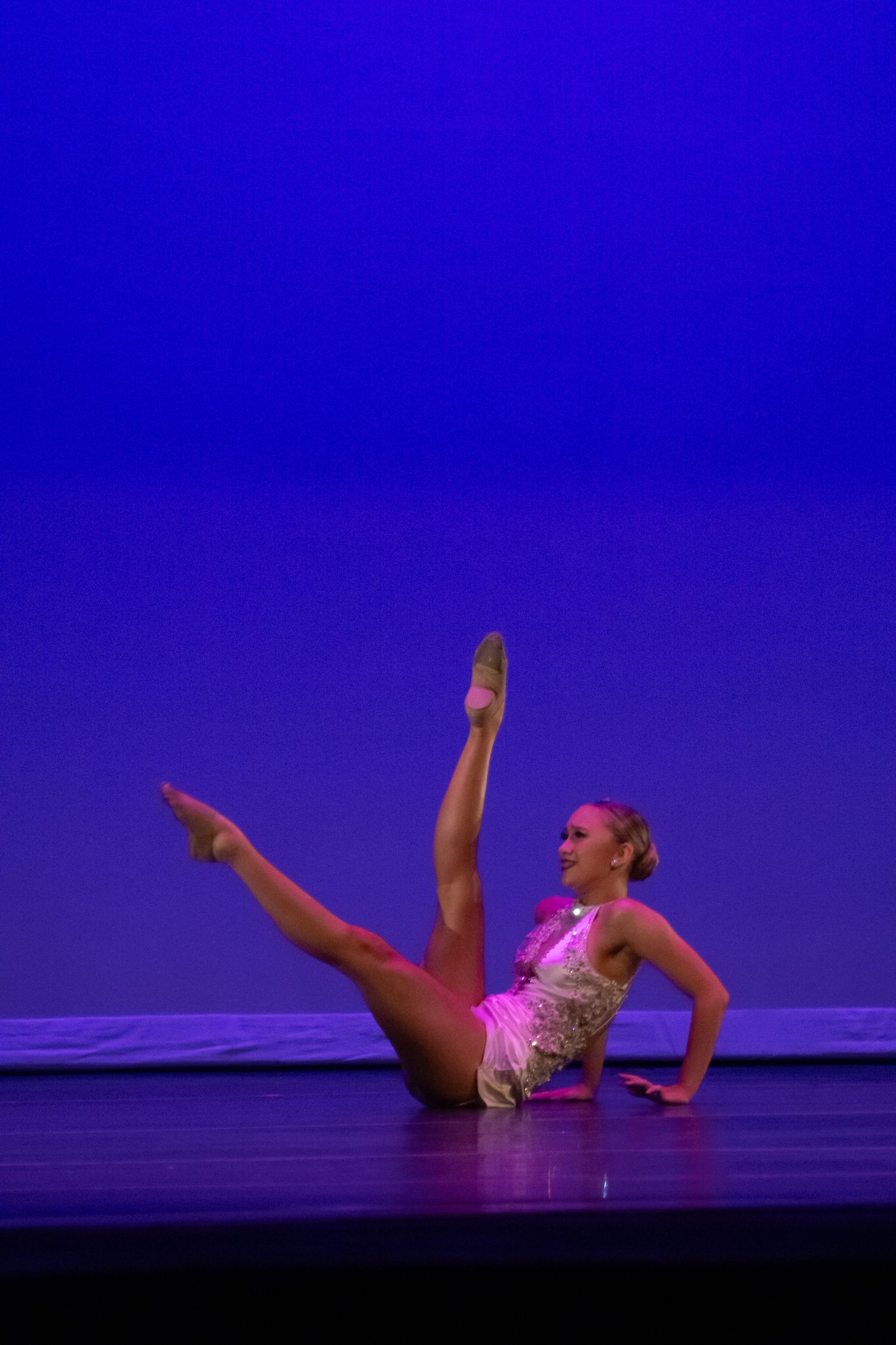 A dancer in a pink leotard performs a floor routine with a high leg extension under blue stage lights.