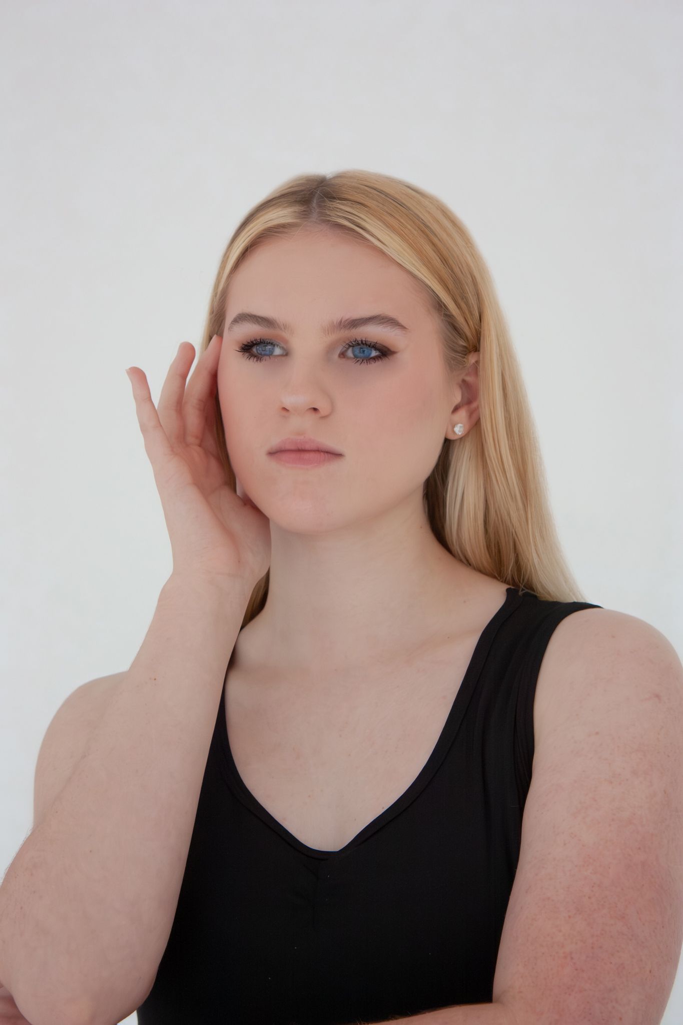 Blonde blue-eyed woman in black tank top gently touches her face against a white background.