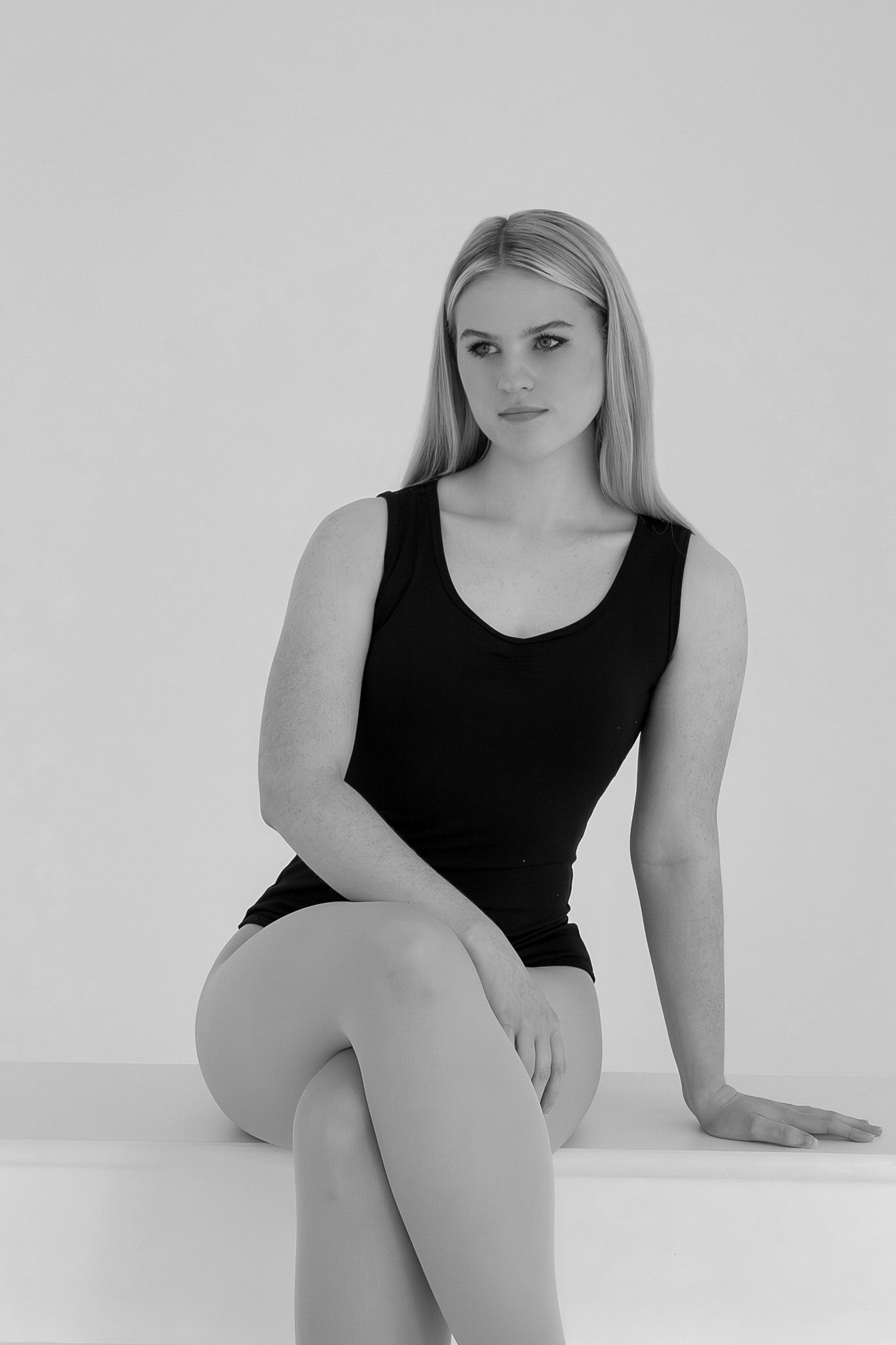 Black and white portrait of a blonde woman in a black leotard sitting on a white surface.