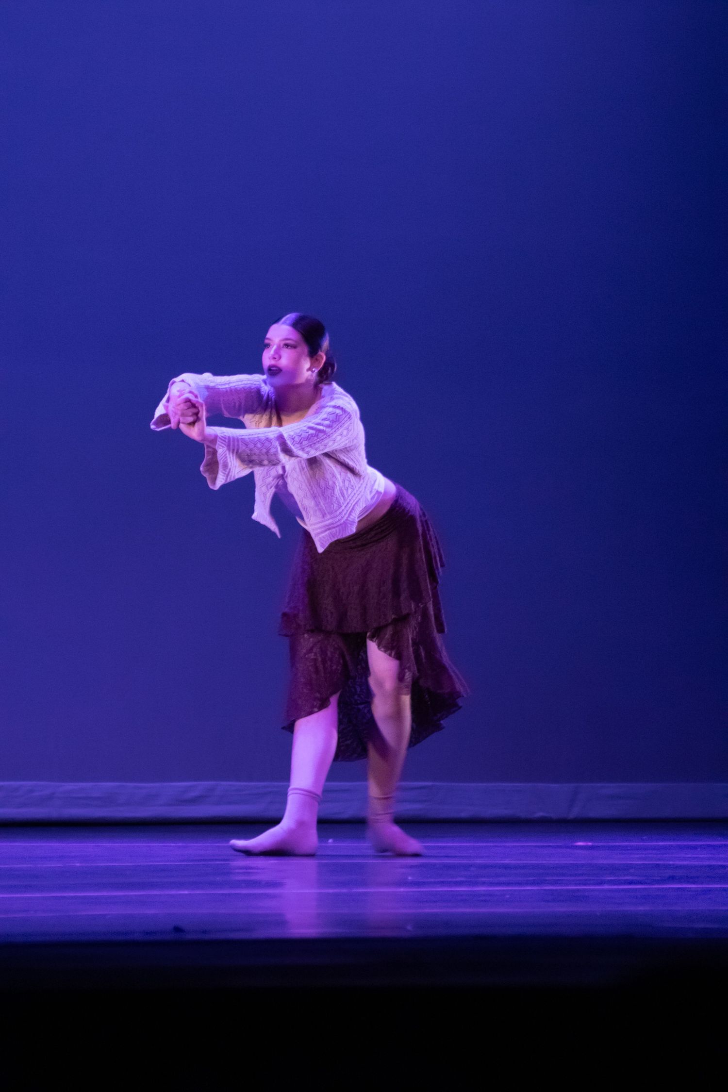 A female dancer in a white top and brown skirt performs a dramatic contemporary piece on a blue-lit stage.