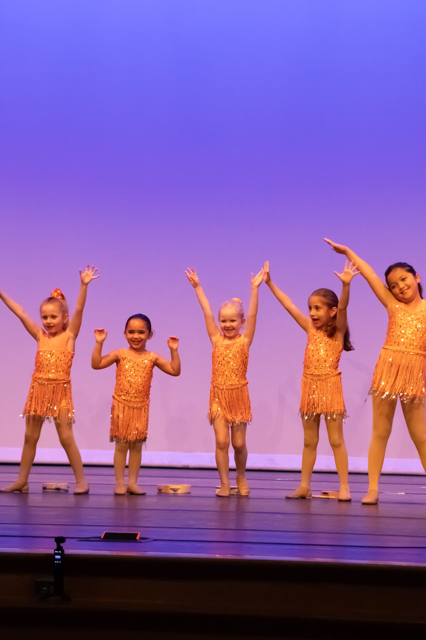 Five young girls in orange sequin fringe costumes raise arms joyfully on a purple-lit stage.