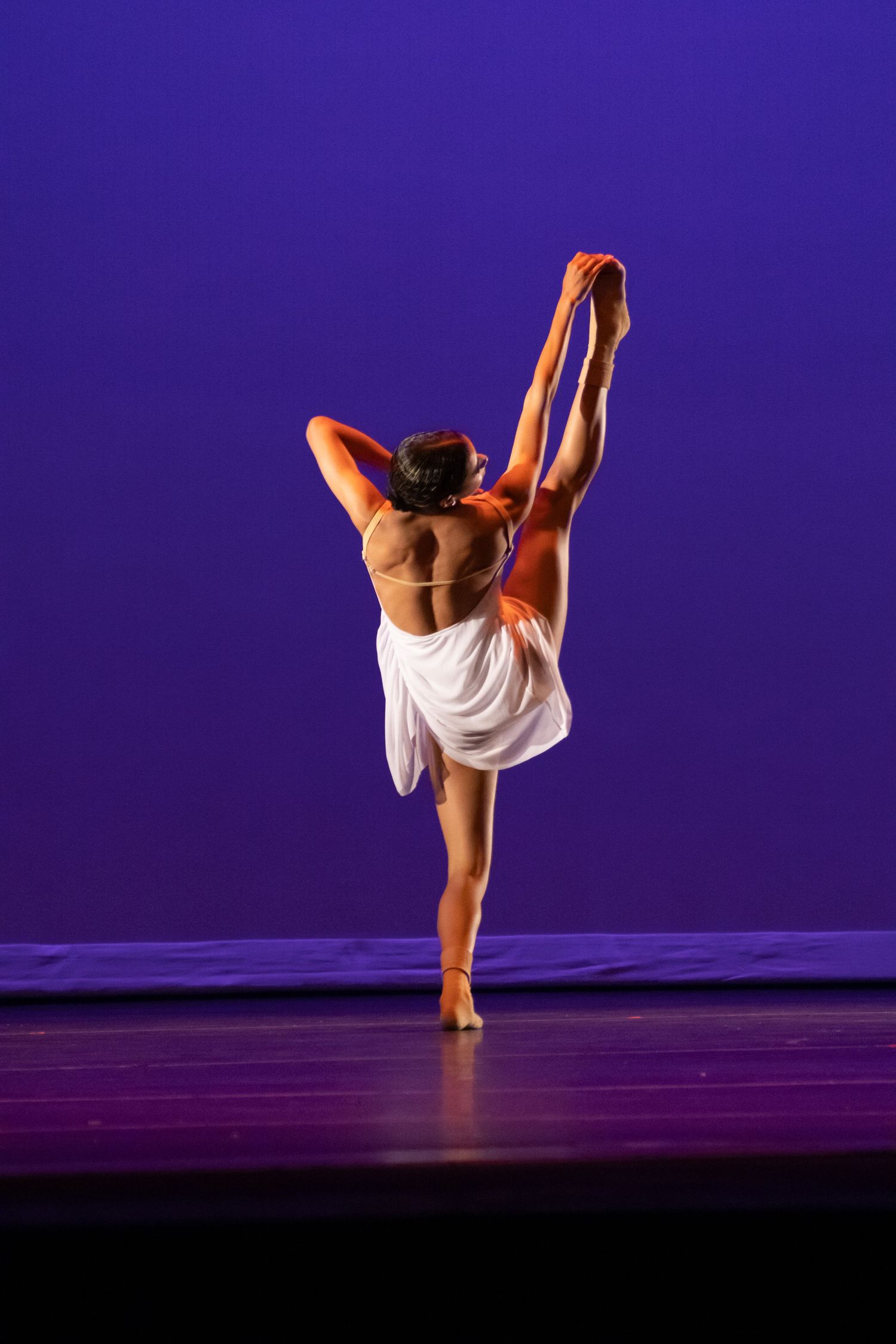 A dancer in white holds a high leg extension from behind on a purple-lit performance stage.