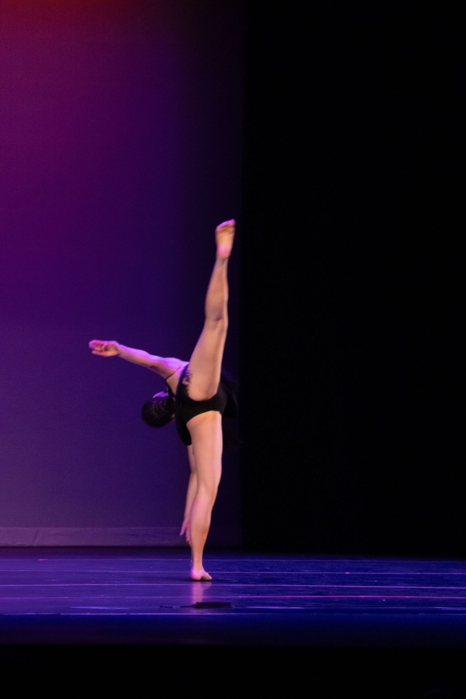 A solo dancer performs a stunning vertical leg extension on stage in a black leotard under purple stage lighting.