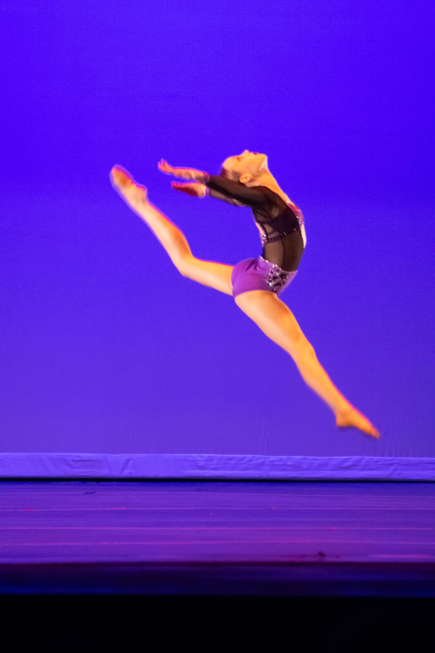 A dancer captures a dynamic leap in a black and purple costume against a vibrant blue stage backdrop.