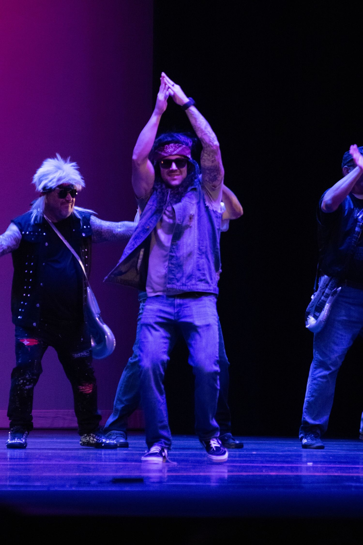 Three performers dance on stage in rock-themed costumes with purple lighting during a live theatrical performance.