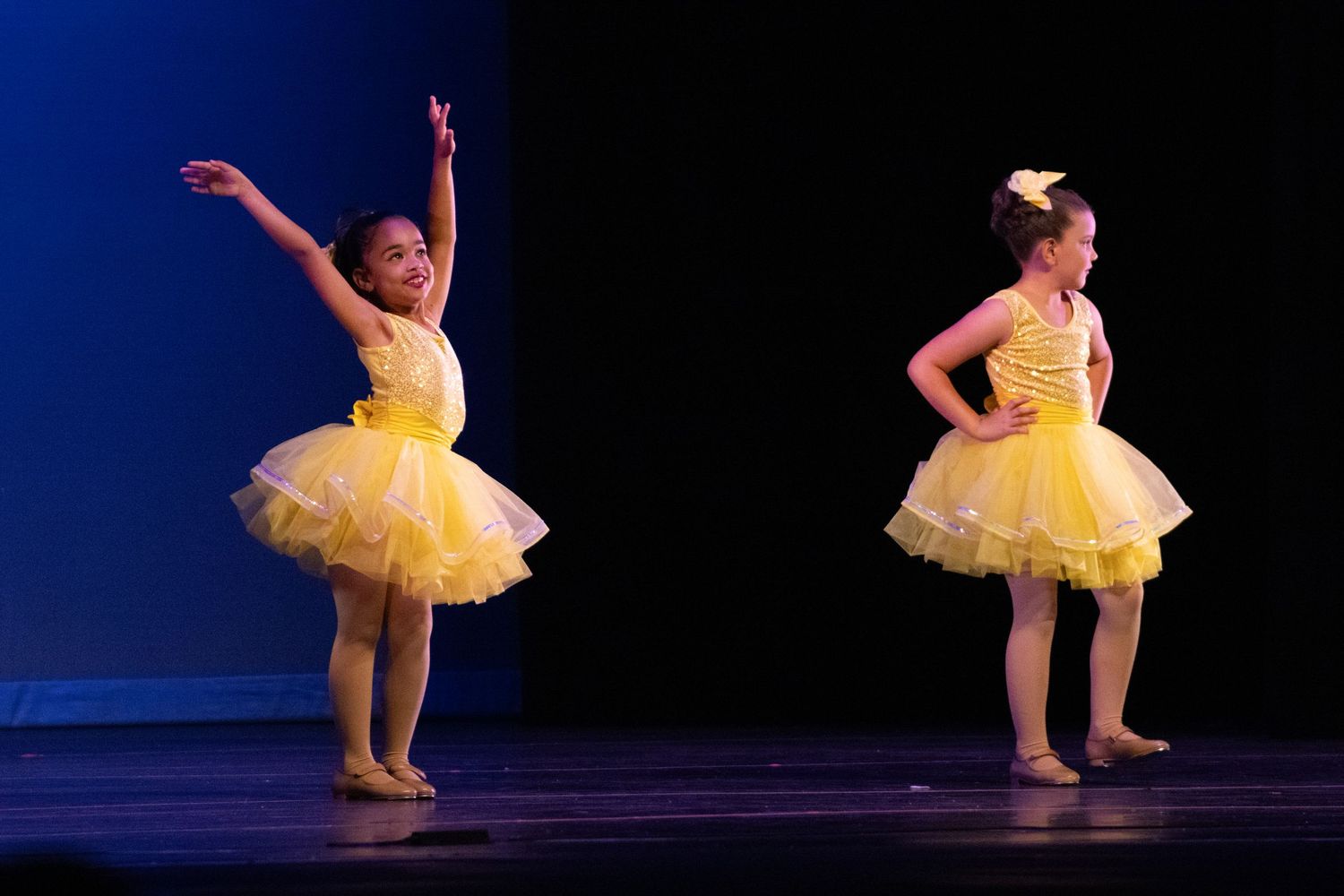 Two young girls in yellow tutus perform ballet on stage during a dance recital.