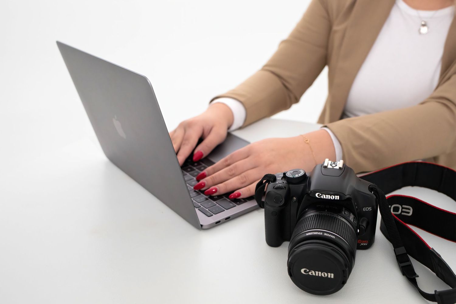 Woman with red nails typing on MacBook laptop next to Canon DSLR camera on white desk.
