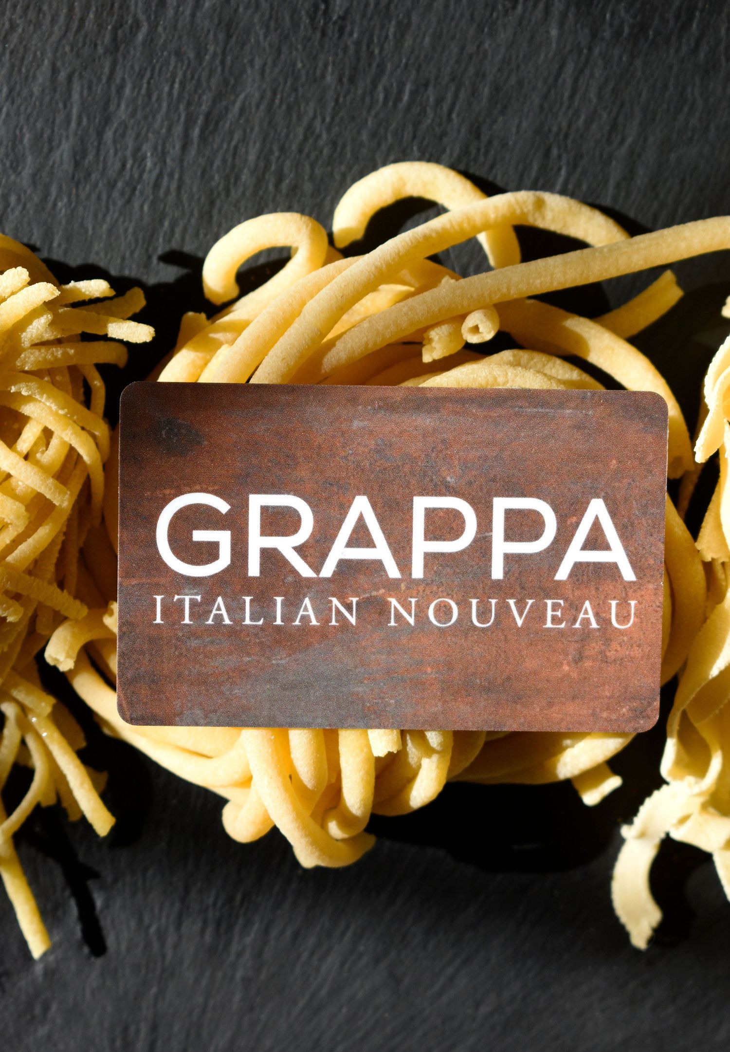 Grappa Italian Nouveau branded card resting on fresh uncooked pasta nests on dark slate surface.