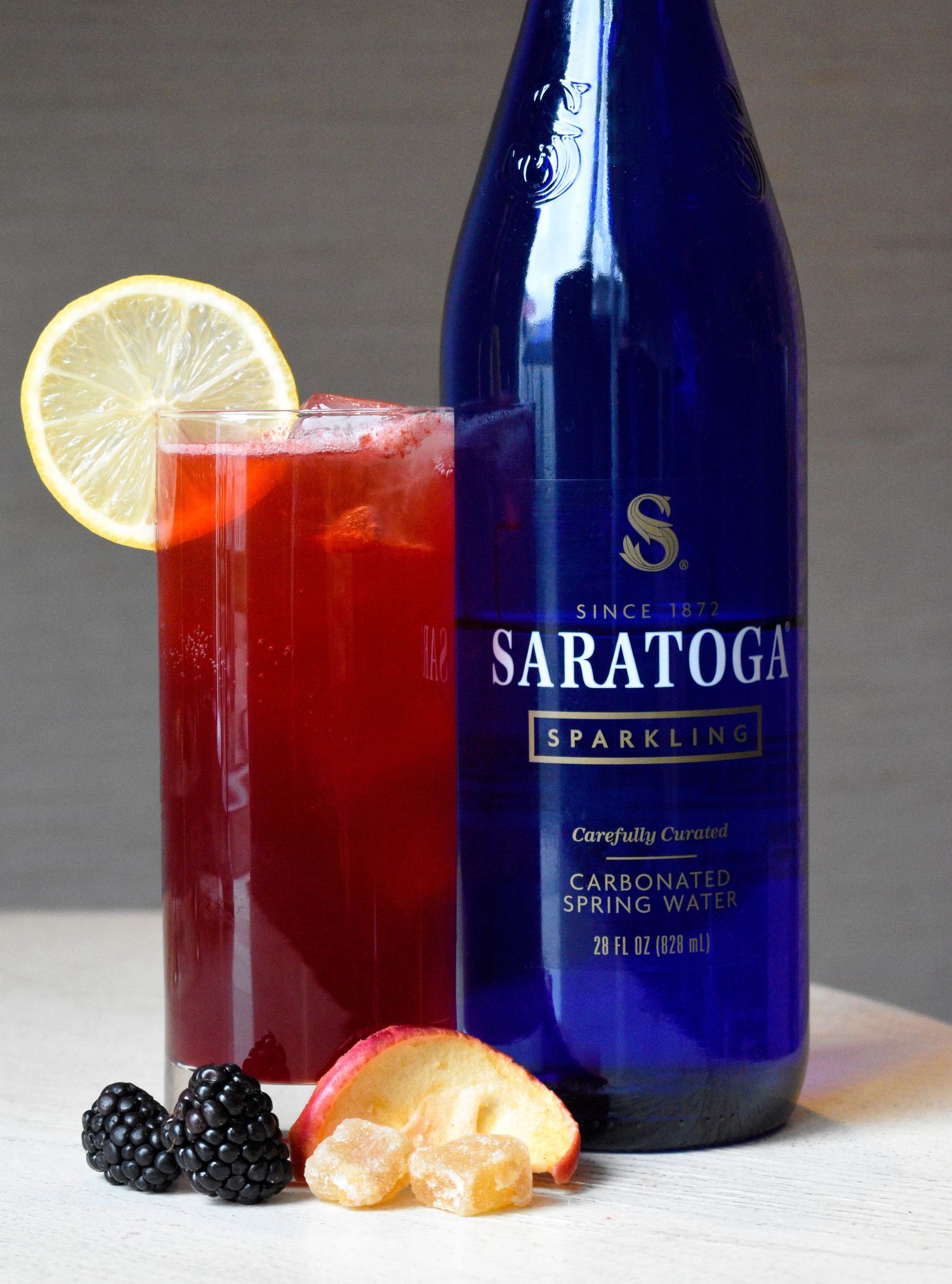Red berry cocktail with lemon garnish beside a blue Saratoga sparkling water bottle with fresh blackberries and peach slices.