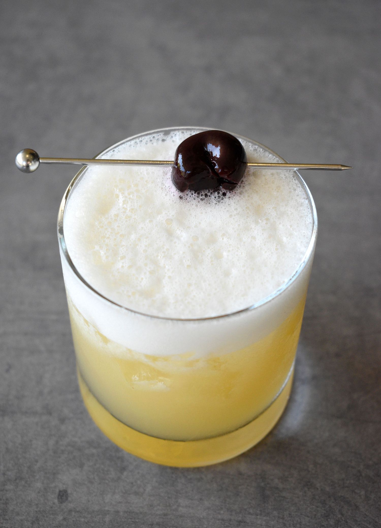 Whiskey sour cocktail with frothy egg white foam and dark cherry garnish on a metal pick.