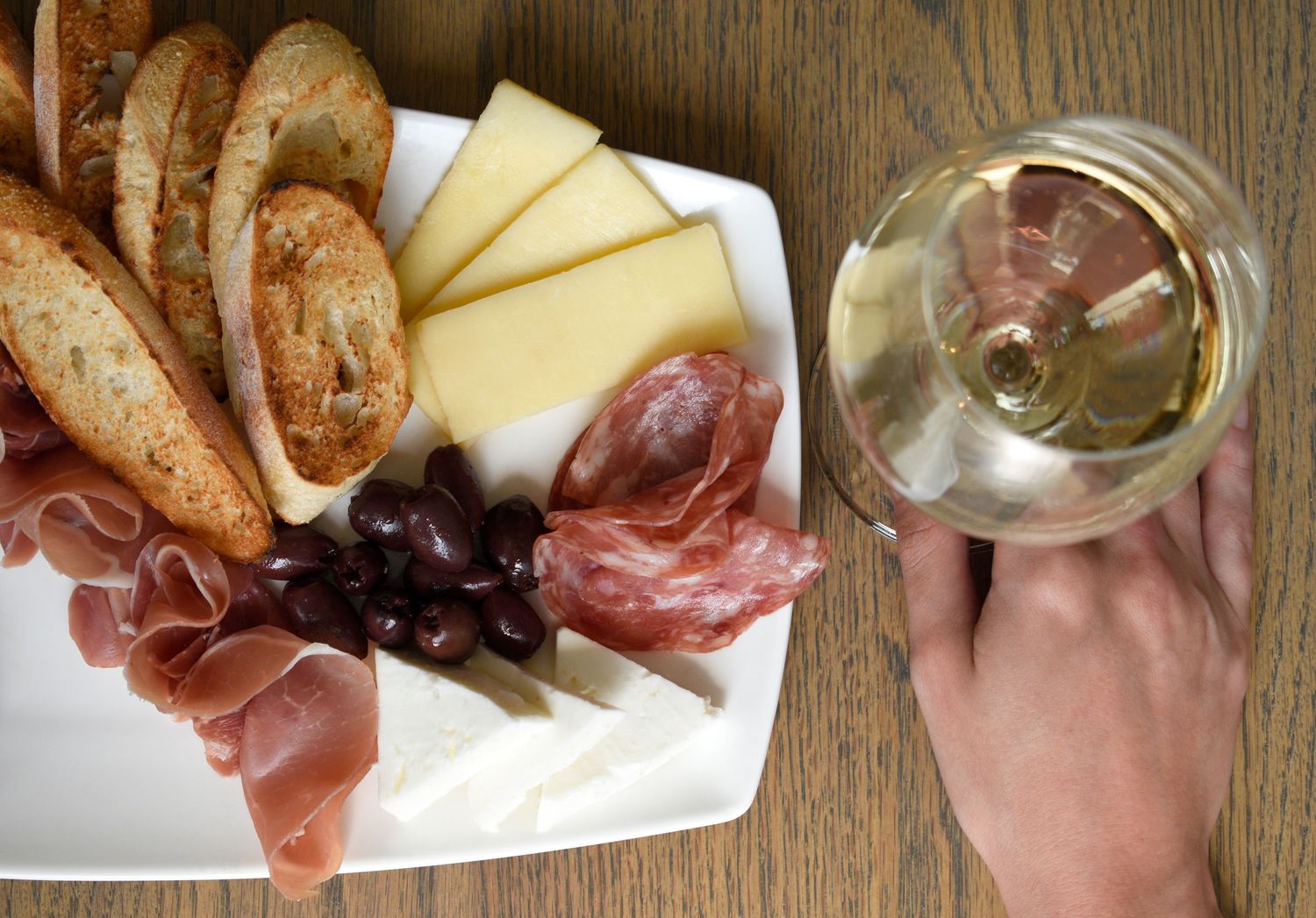 Hand holding white wine glass beside charcuterie board with meats, cheese, olives, and toasted baguette slices.