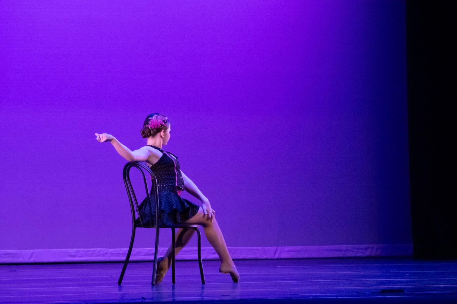 Young dancer in dark costume performs chair routine under purple stage lighting.