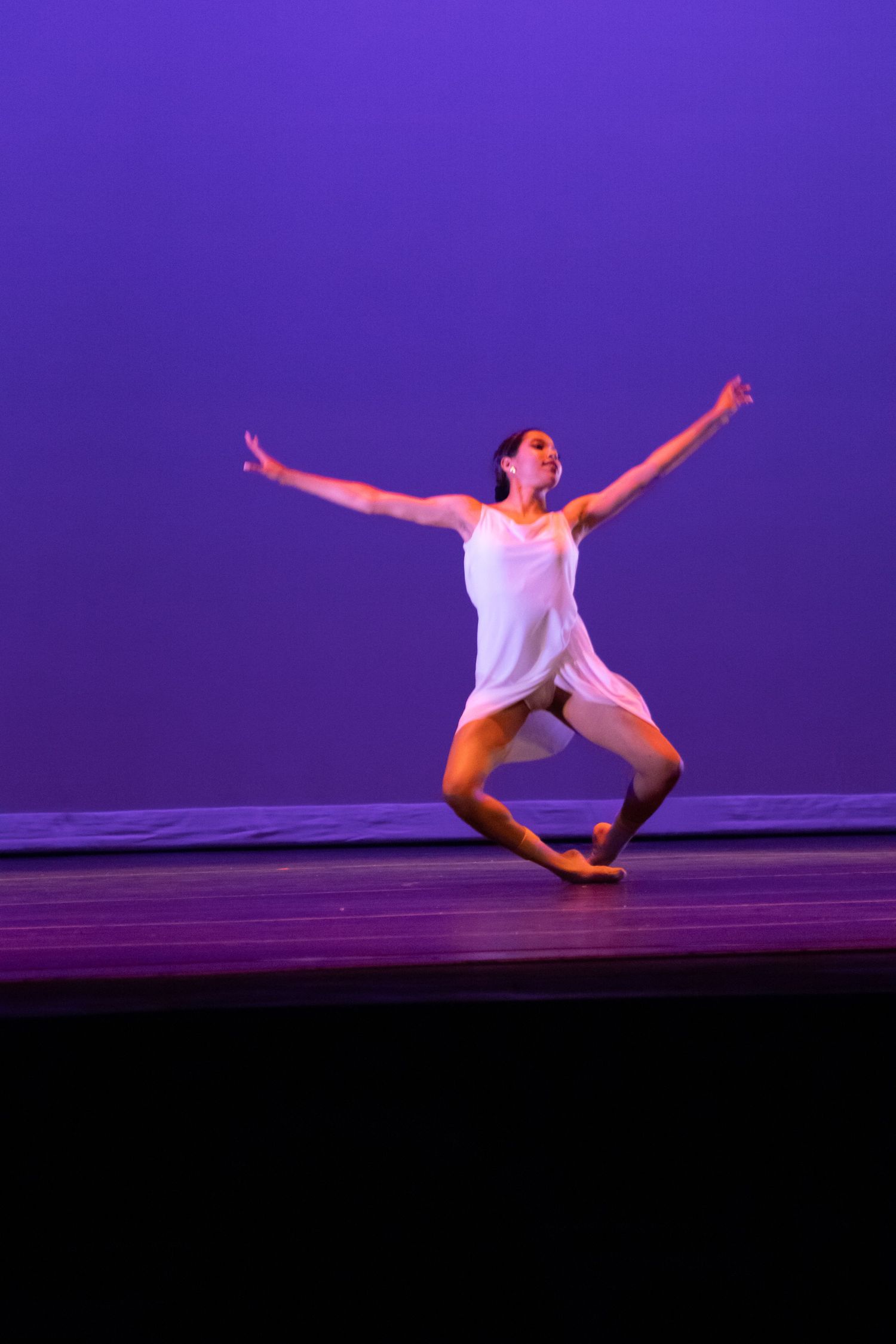 Female dancer in white dress performs expressive contemporary solo on purple-lit stage.