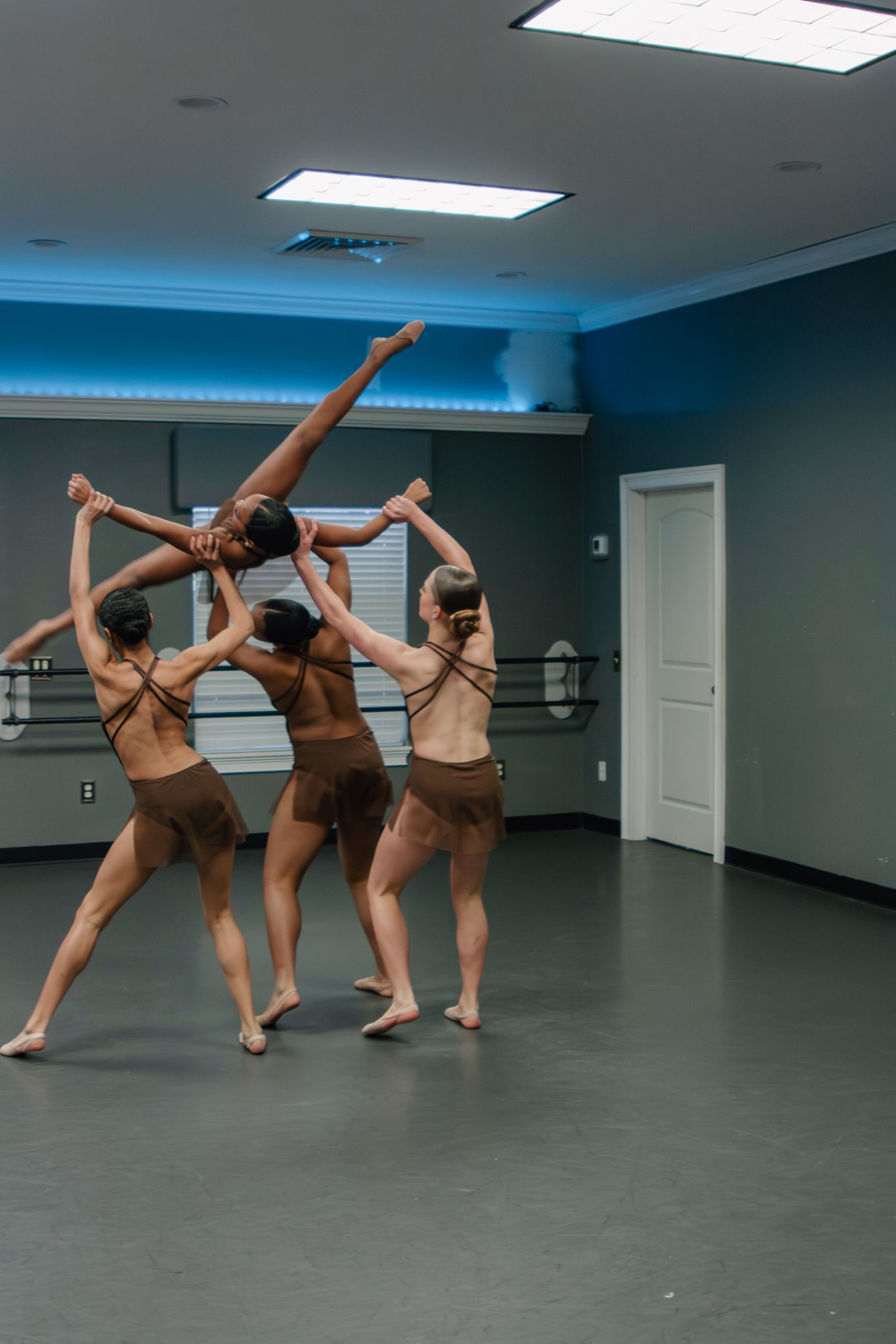 Four contemporary dancers in brown leotards perform a group lift routine in a mirrored dance studio.