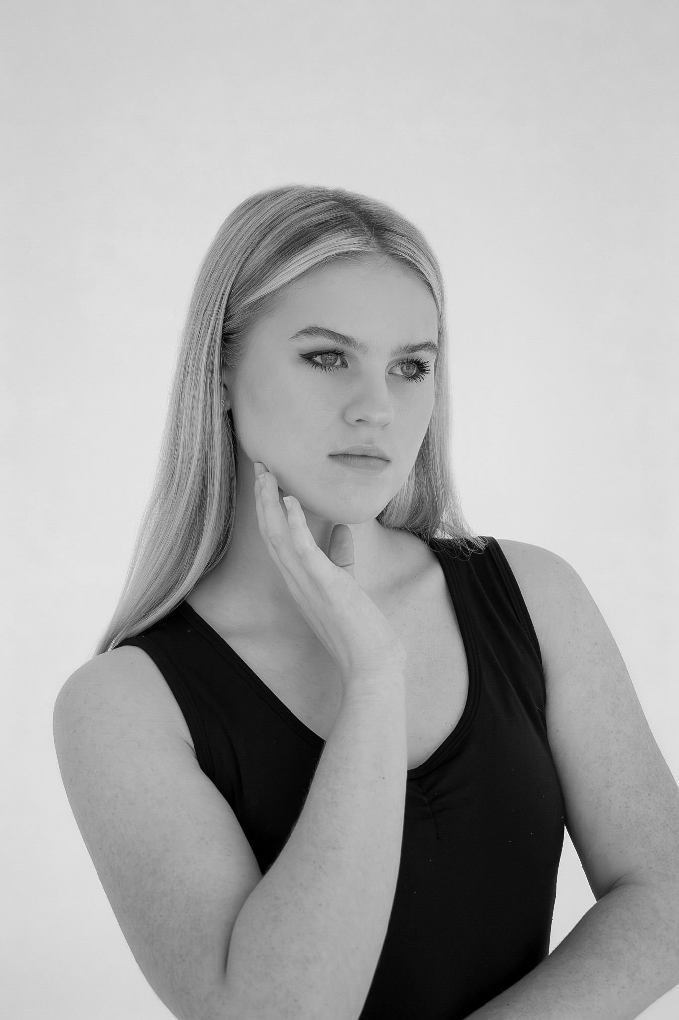 Black and white portrait of a young blonde woman in a black top, hand on cheek, against a light background.