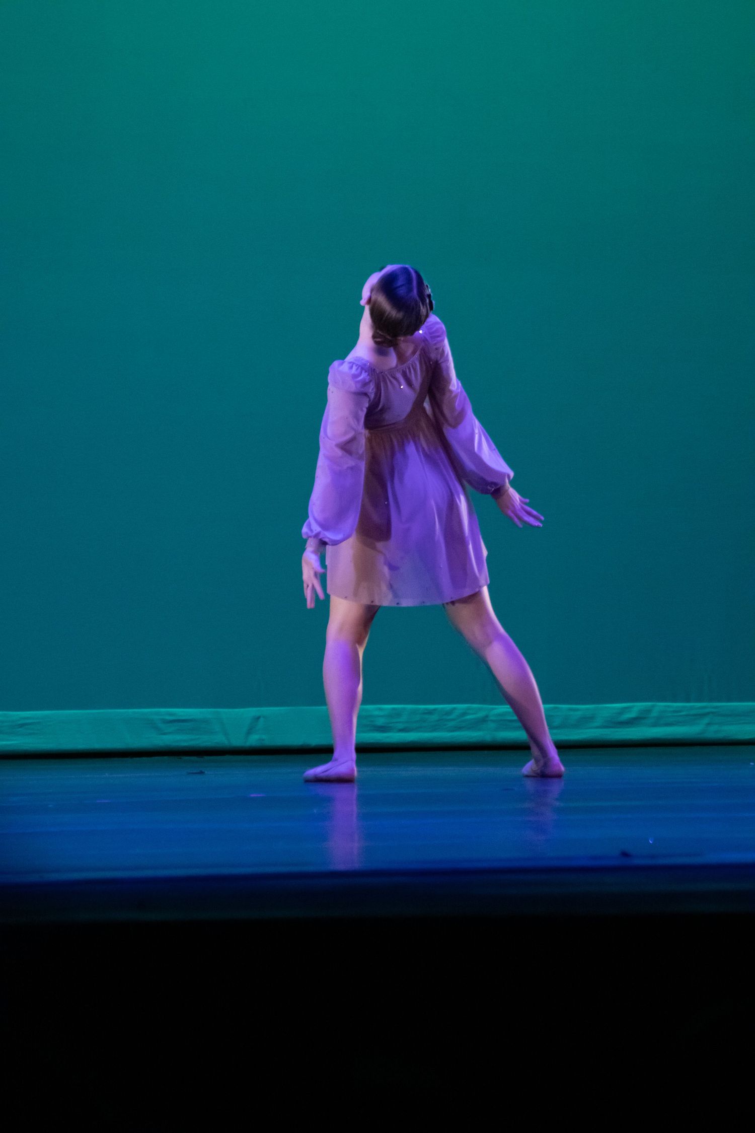 A solo dancer in a flowing lavender dress performs a contemporary dance routine on a teal-lit stage.