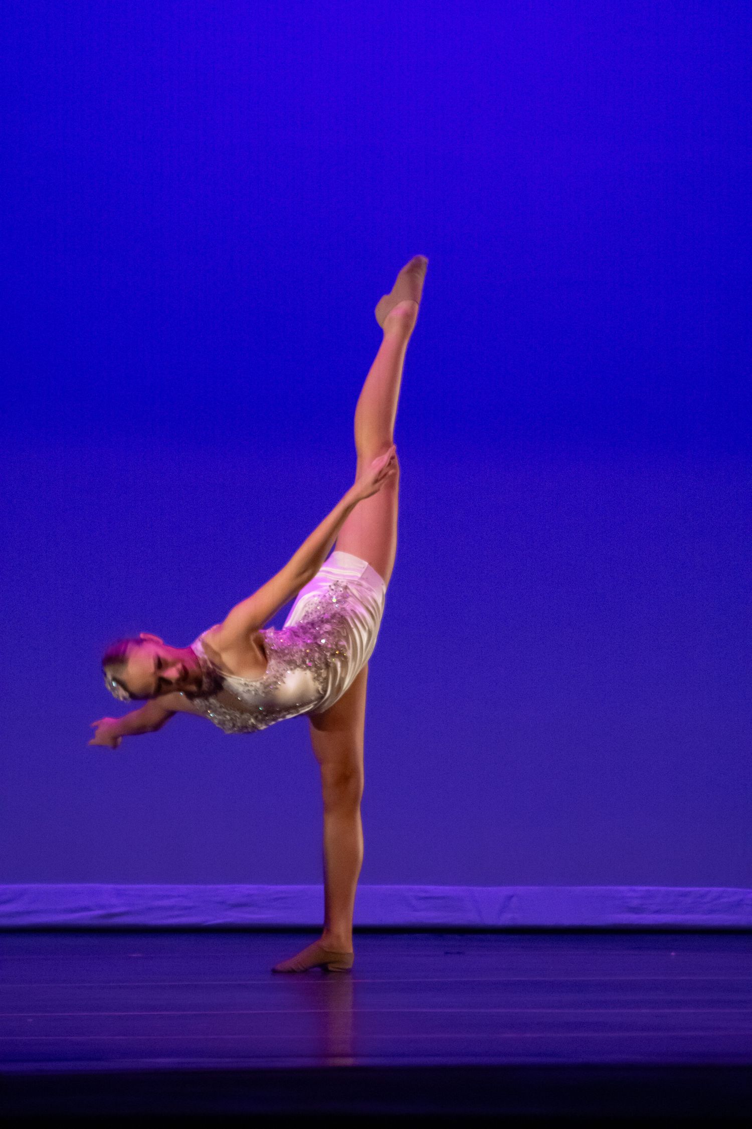 A skilled dancer performs a dramatic needle balance pose in a pink and white costume under blue stage lights.