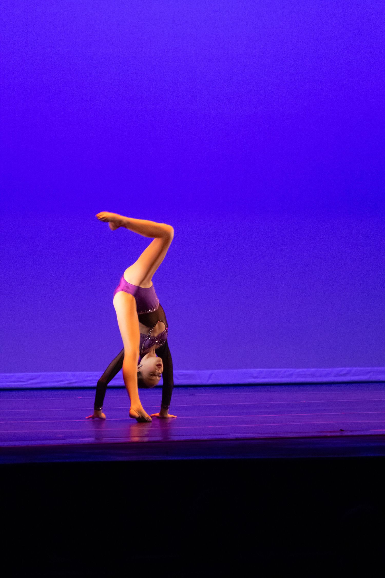 A young dancer performs a handstand with bent knees on a blue-lit stage in a purple costume.