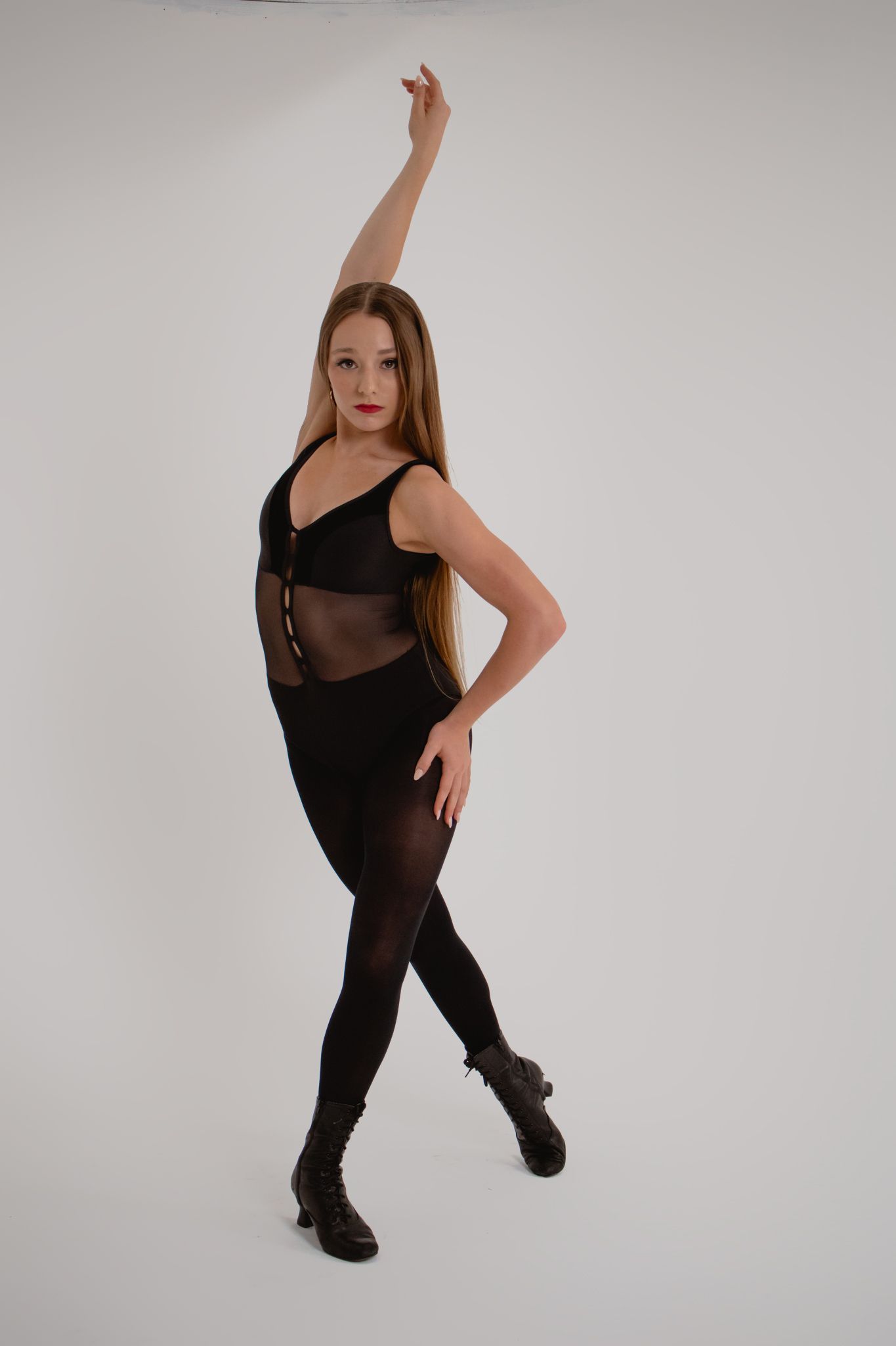 Female dancer in black bodysuit and boots raises arm in elegant pose against white background.