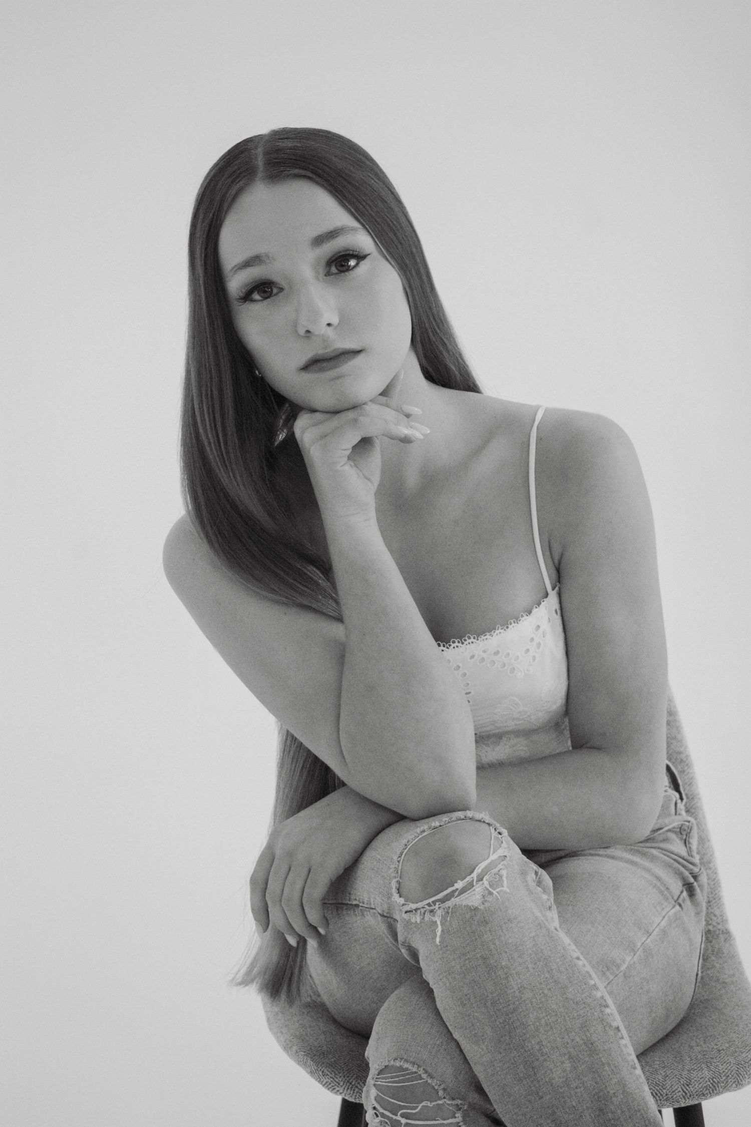 Black and white portrait of a young woman with long hair sitting pensively with hand on chin.