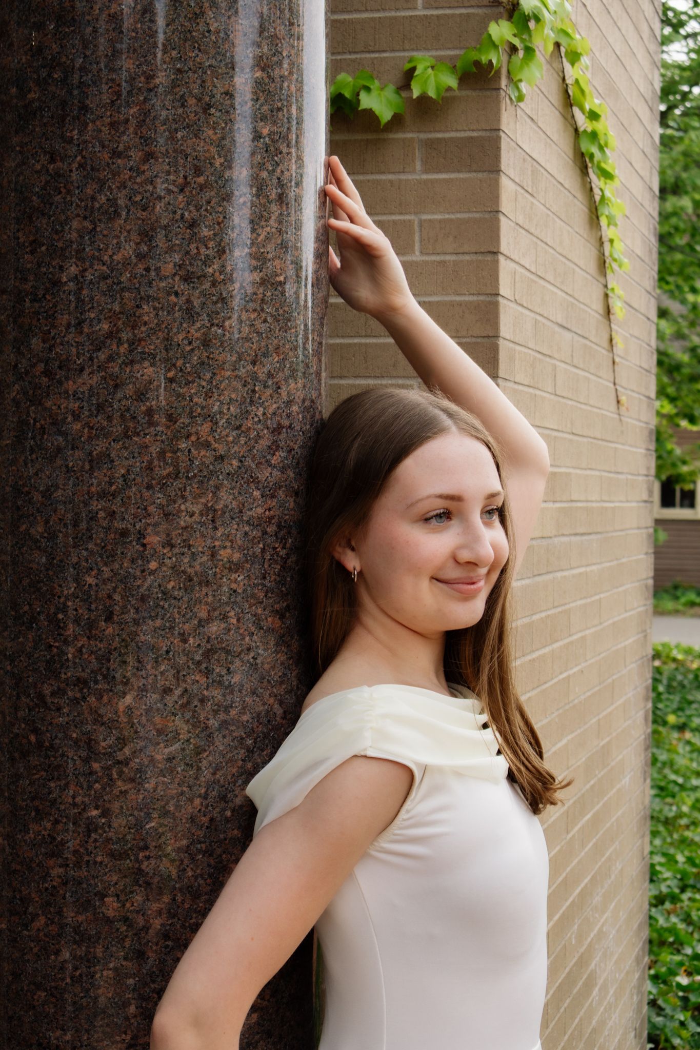 Young woman in white off-shoulder dress leans gracefully against granite column near brick wall.