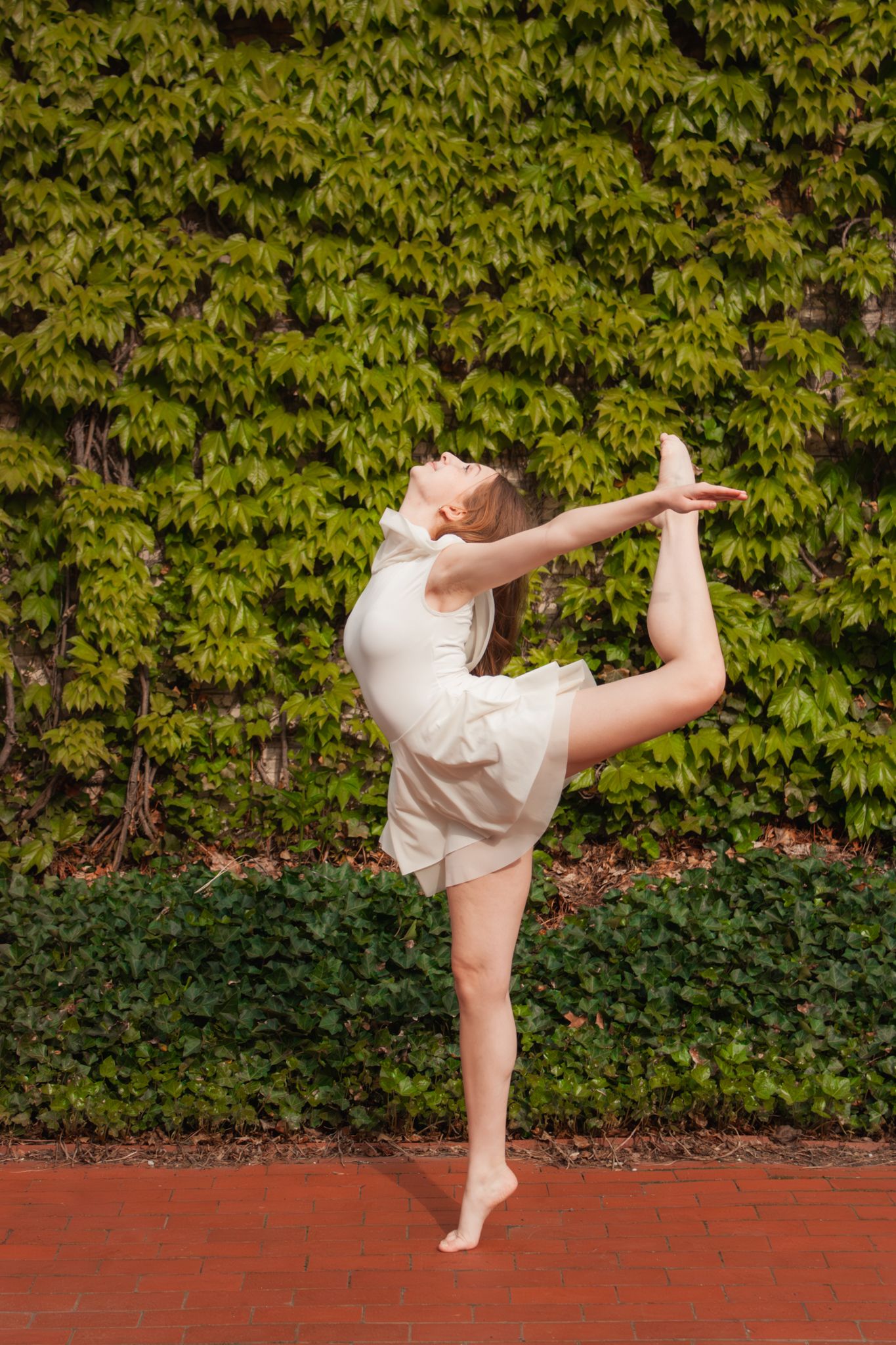 A flexible ballerina performs an arabesque pose outdoors in a white outfit against a lush green ivy-covered wall.