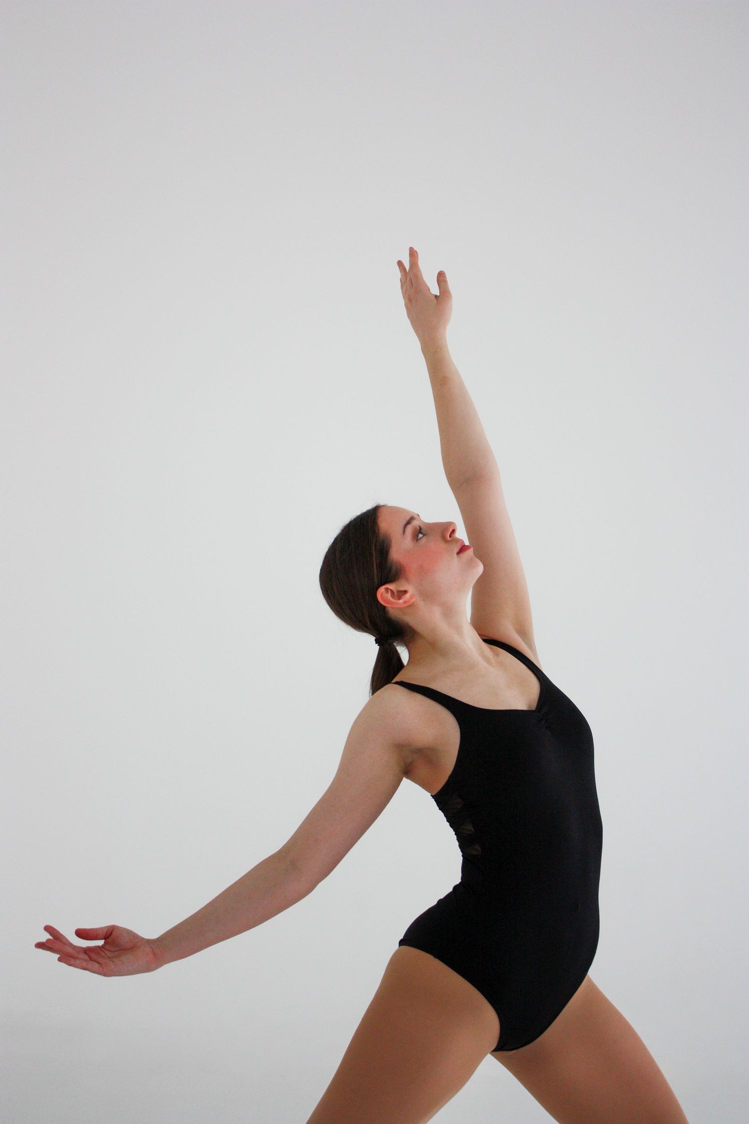 A female dancer in a black leotard stretches one arm upward while arching back gracefully against a white background.