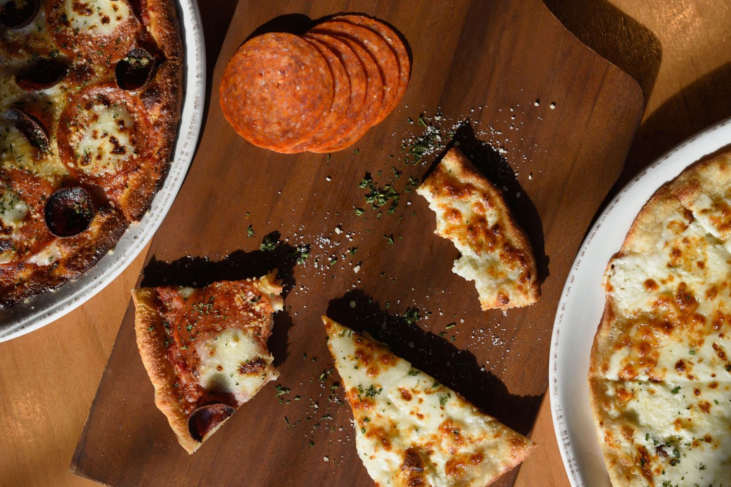 Sliced deep dish and white cheese pizza on a wooden board with pepperoni nearby.