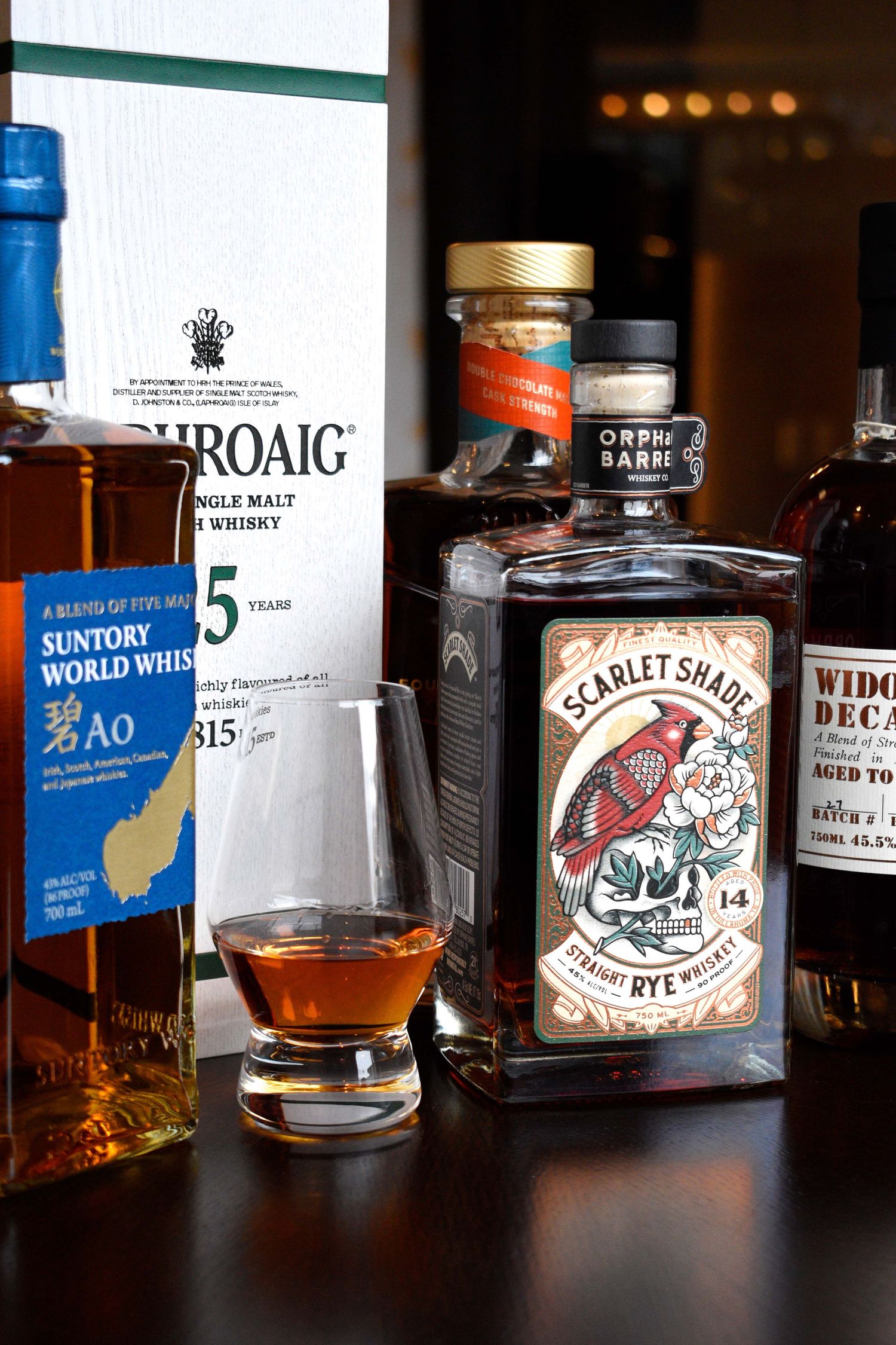 Collection of premium whisky bottles including Scarlet Shade Rye, Laphroaig, and Suntory Ao with a glass of amber whisky.