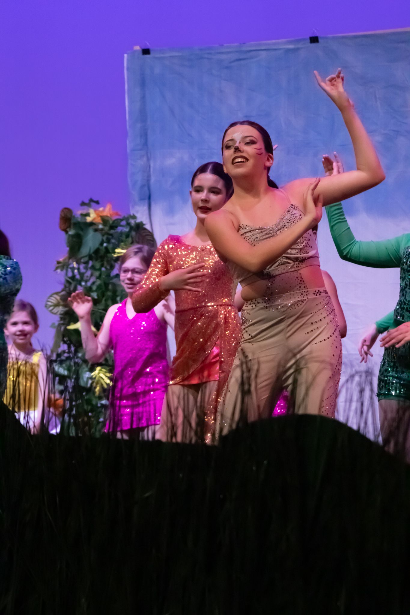 Female dancers in sparkly sequin costumes perform an expressive dance on a purple-lit theatrical stage.