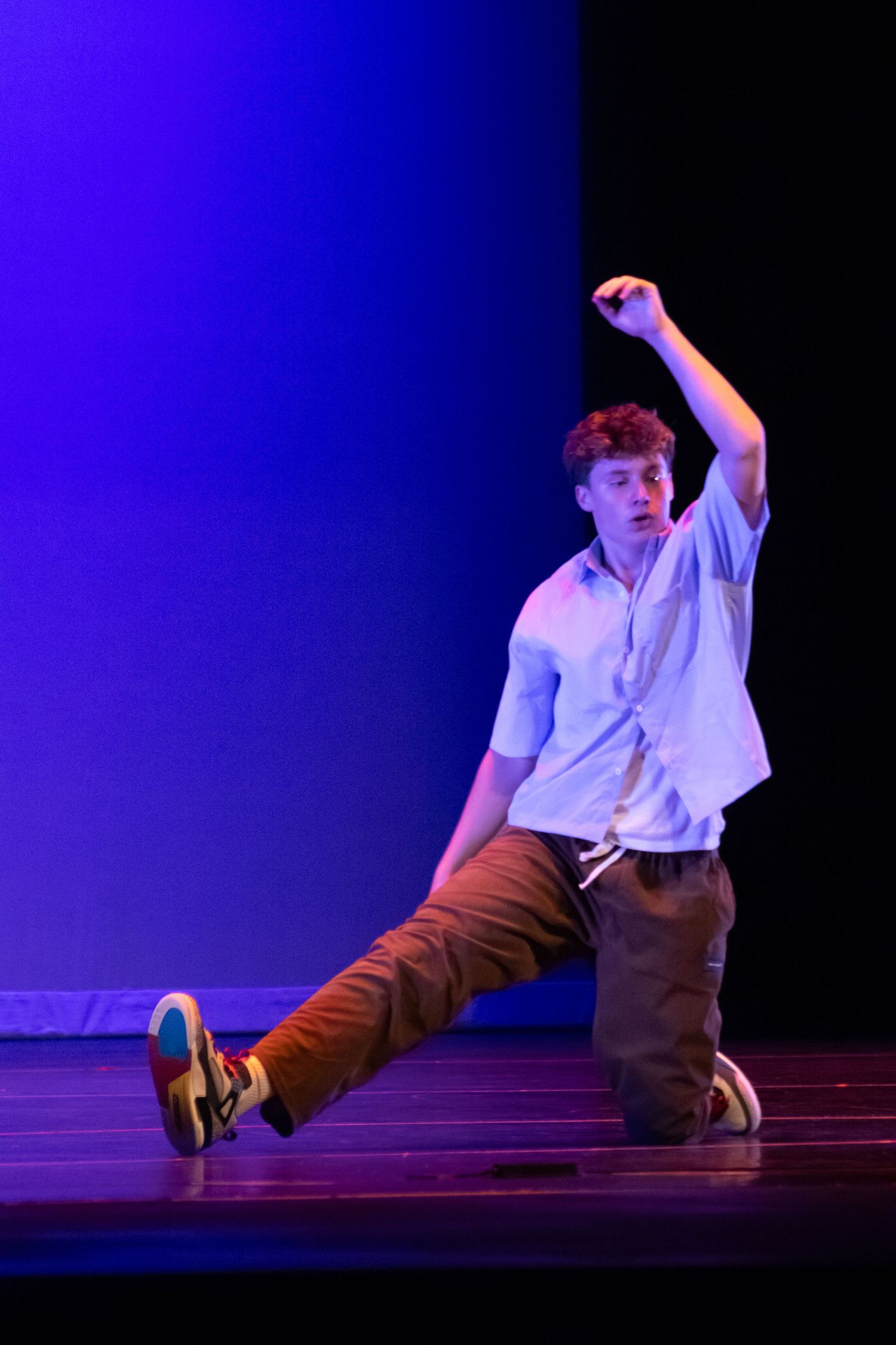 Male dancer in casual clothes performs a dynamic kneeling pose on a blue-lit stage.