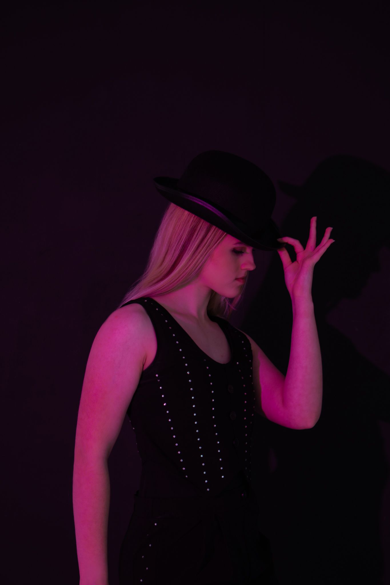 Blonde woman in black hat and studded vest poses under moody pink-purple neon studio lighting.