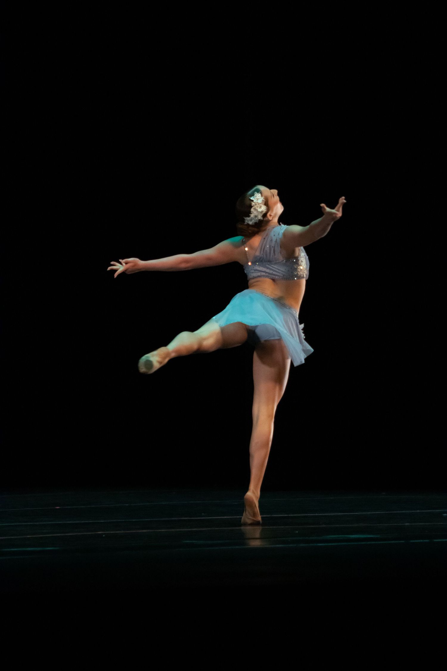 Female ballet dancer in blue tutu performs an arabesque on a dark stage.