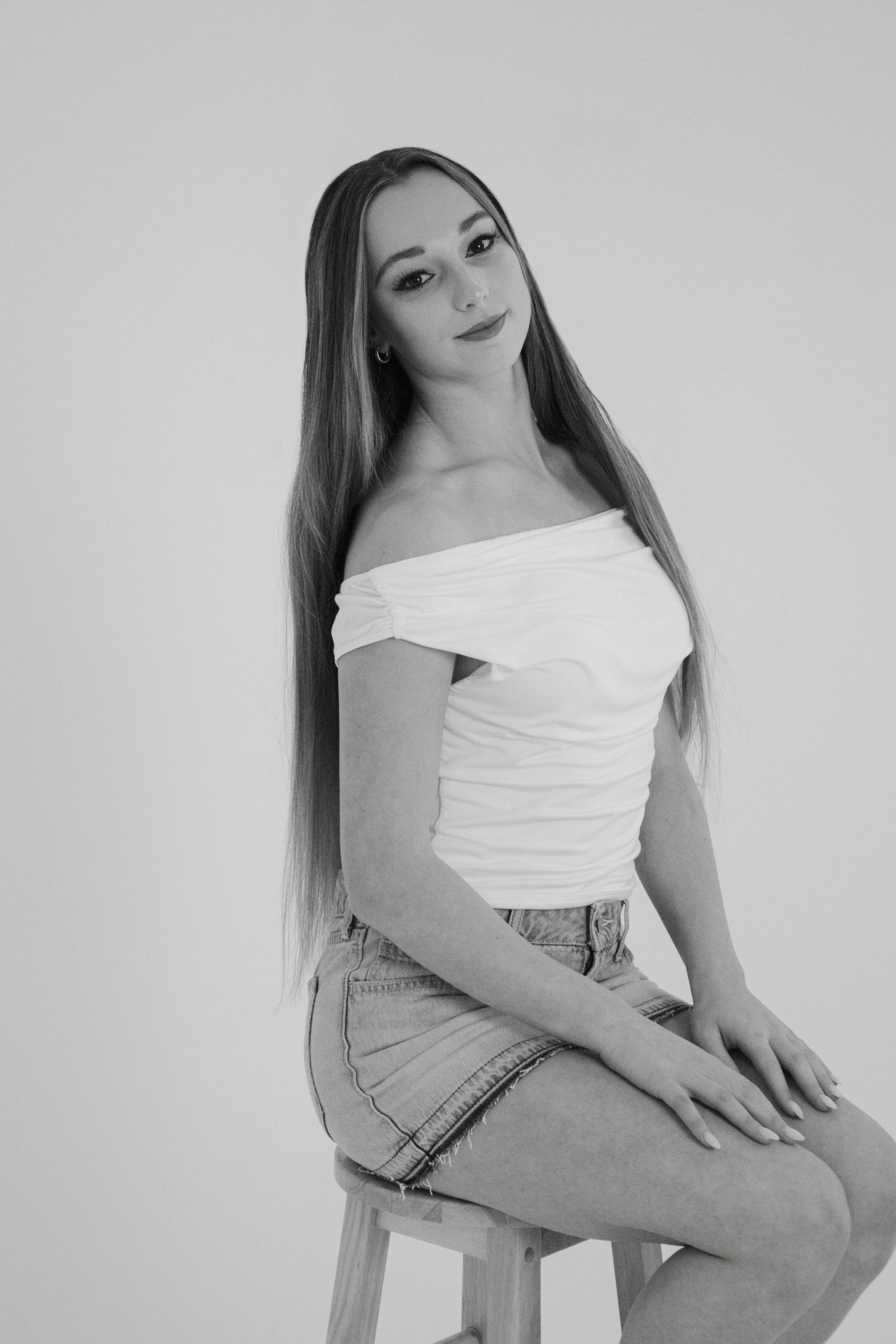 Young woman with long hair poses on stool in off-shoulder white top and denim skirt, black and white studio photo.